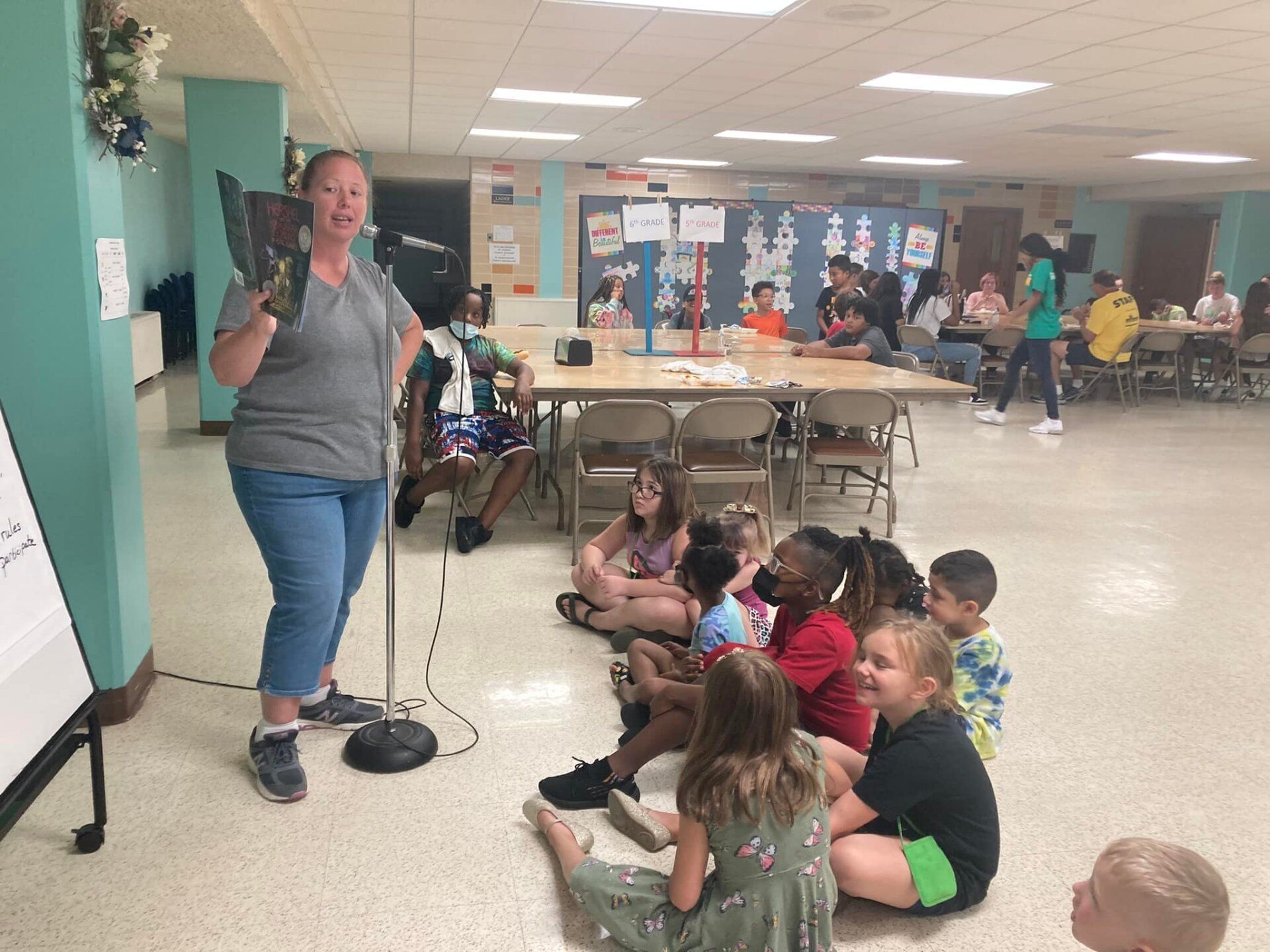 Woman reading a book to children in a community center. Kids sit on the floor, listening.