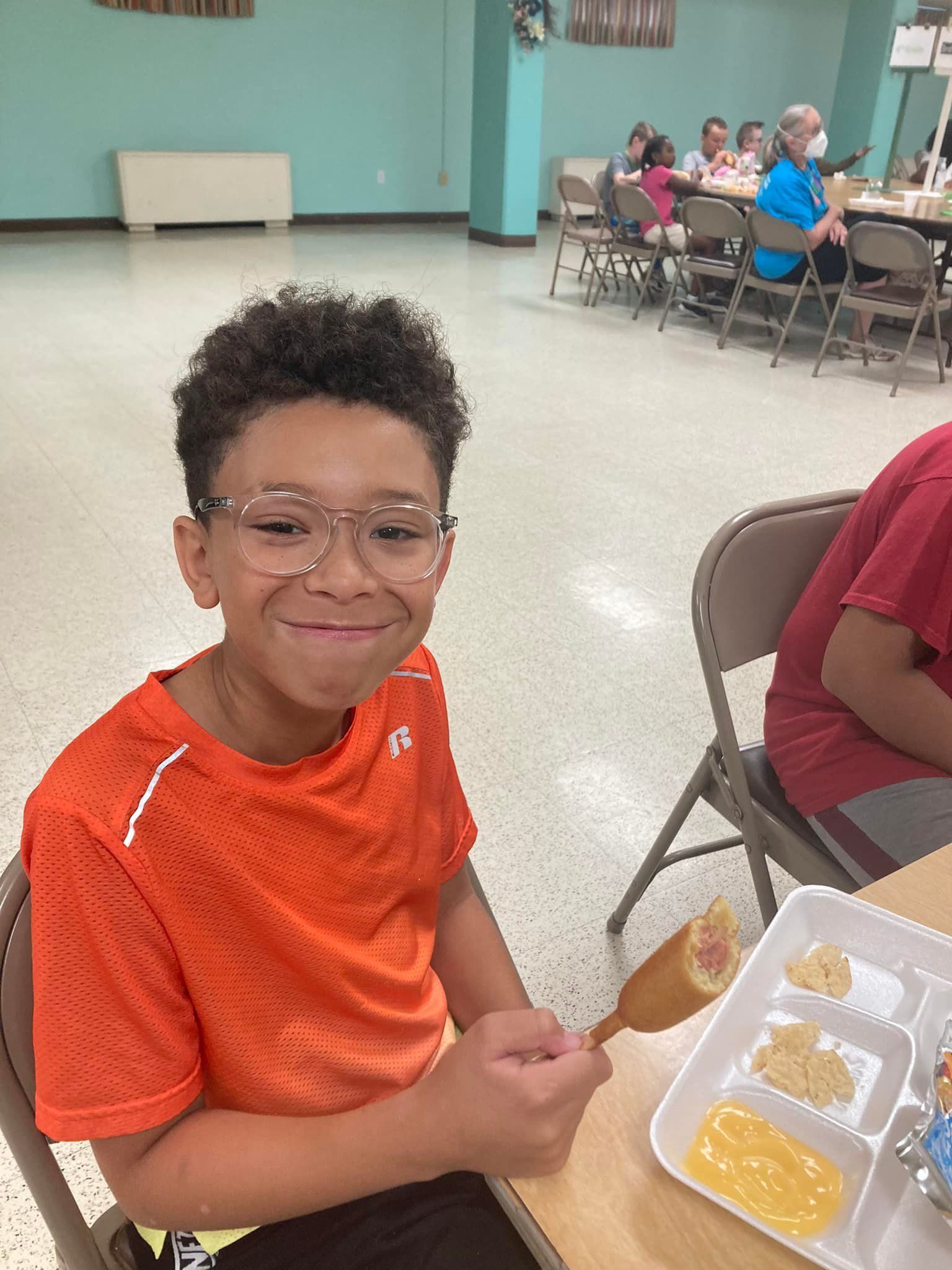 Boy smiles, holds corn dog at lunch table. Orange shirt, glasses, school cafeteria.