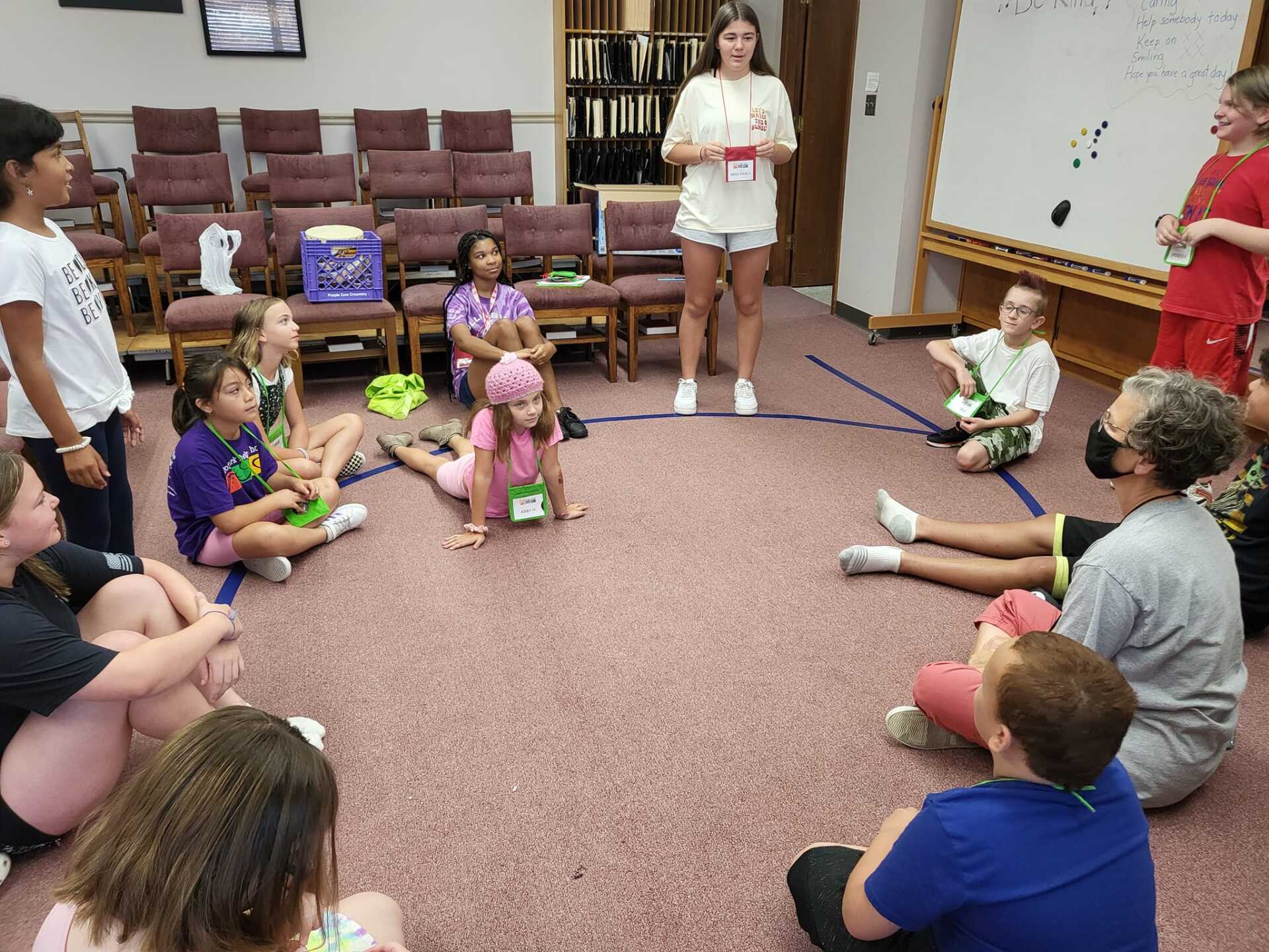 Children in a circle, listening to a girl speak in a library-like room.
