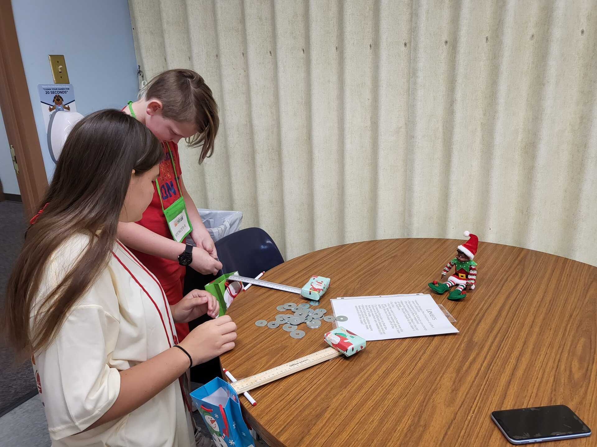 Two kids at a table, possibly playing a game with coins. An elf statue is also on the table.