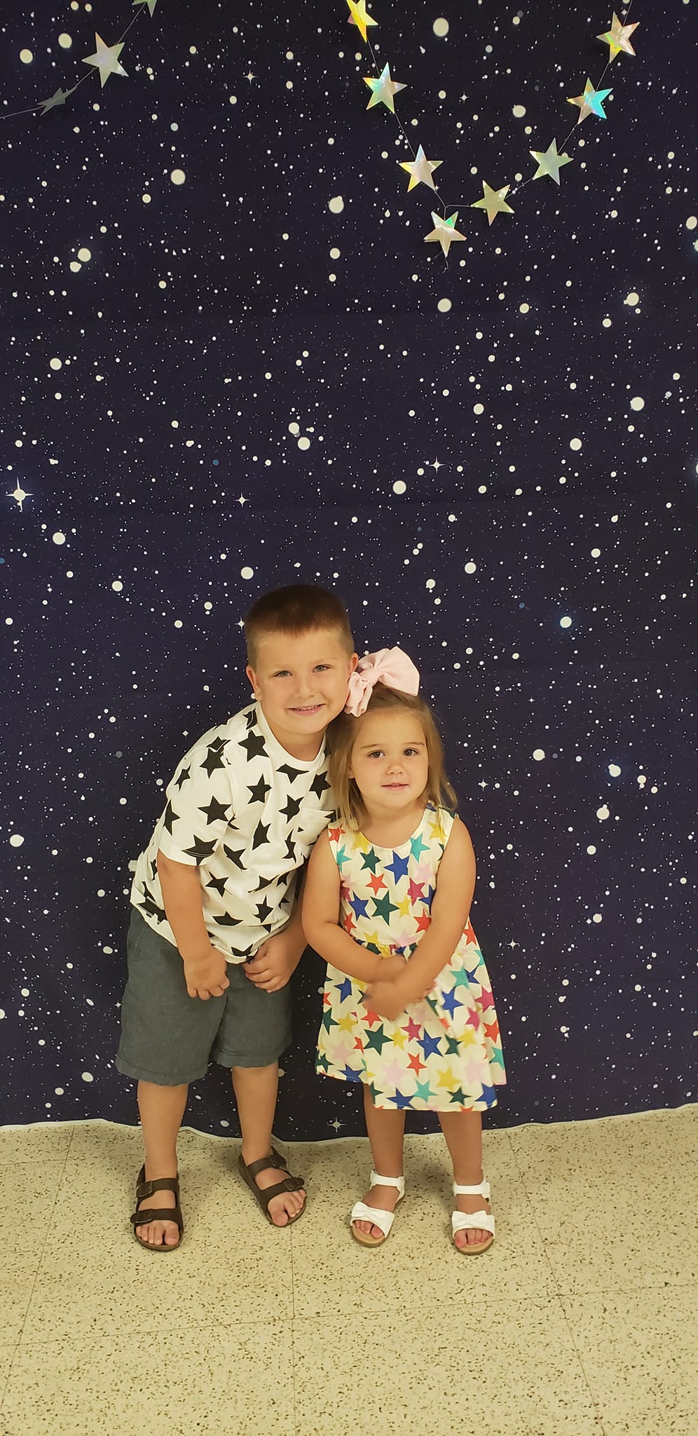 Two young children smile, posing in front of a starry background, boy in shorts, girl in a floral dress.