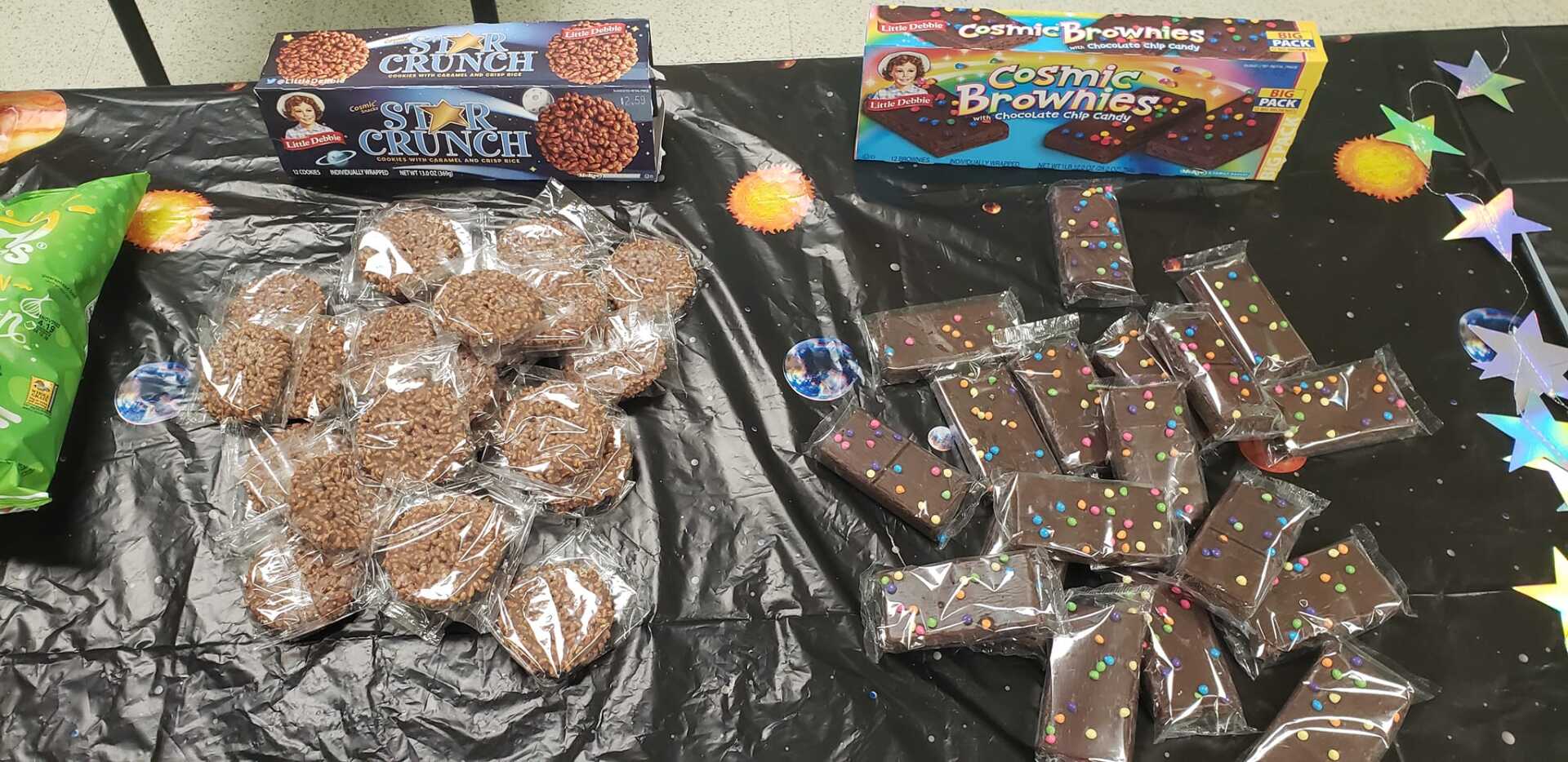 A table displays packaged baked goods, including cookies and brownies, with space-themed decorations.