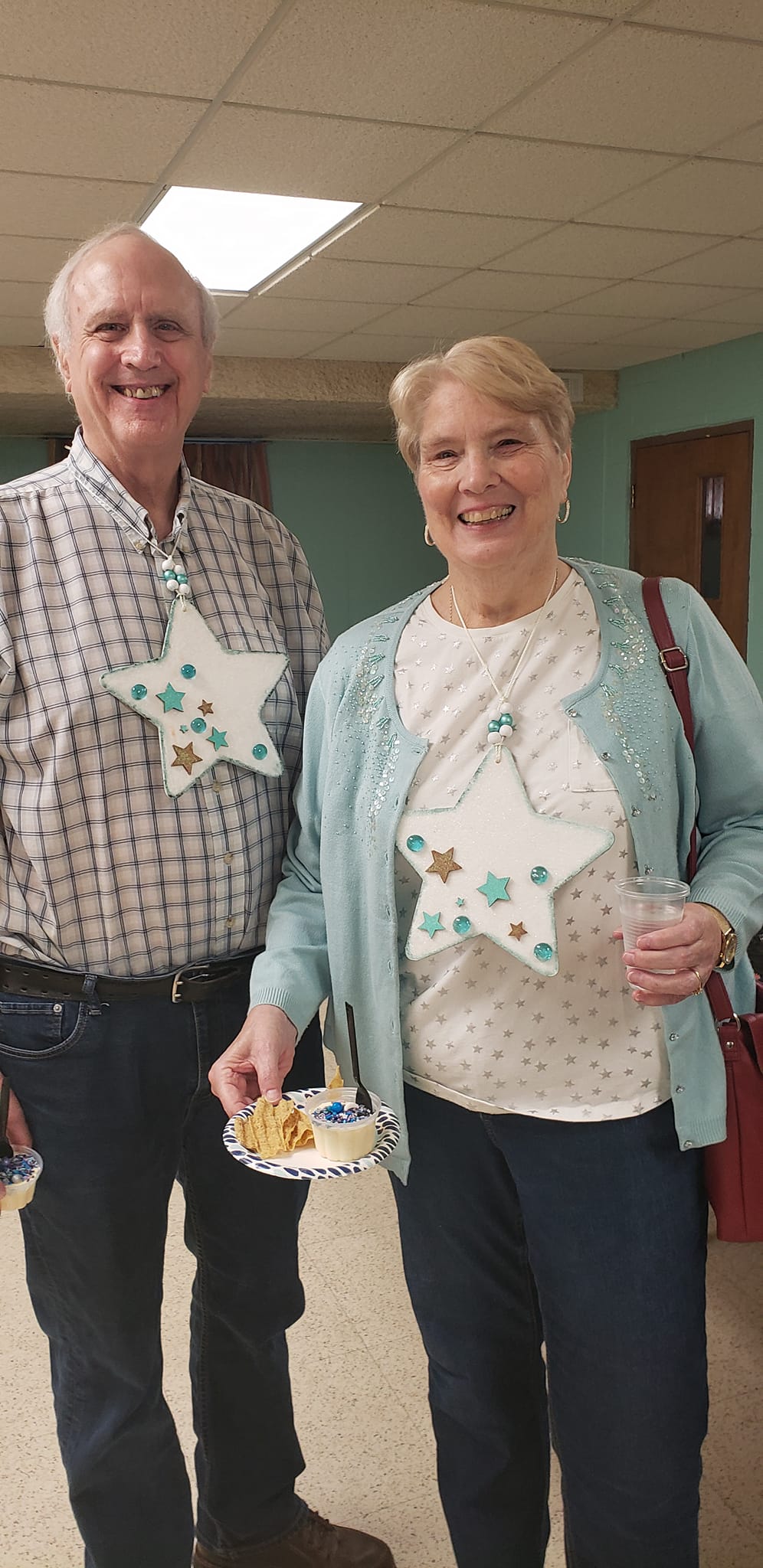 Two people smiling, wearing star-shaped badges and holding food/drink indoors.