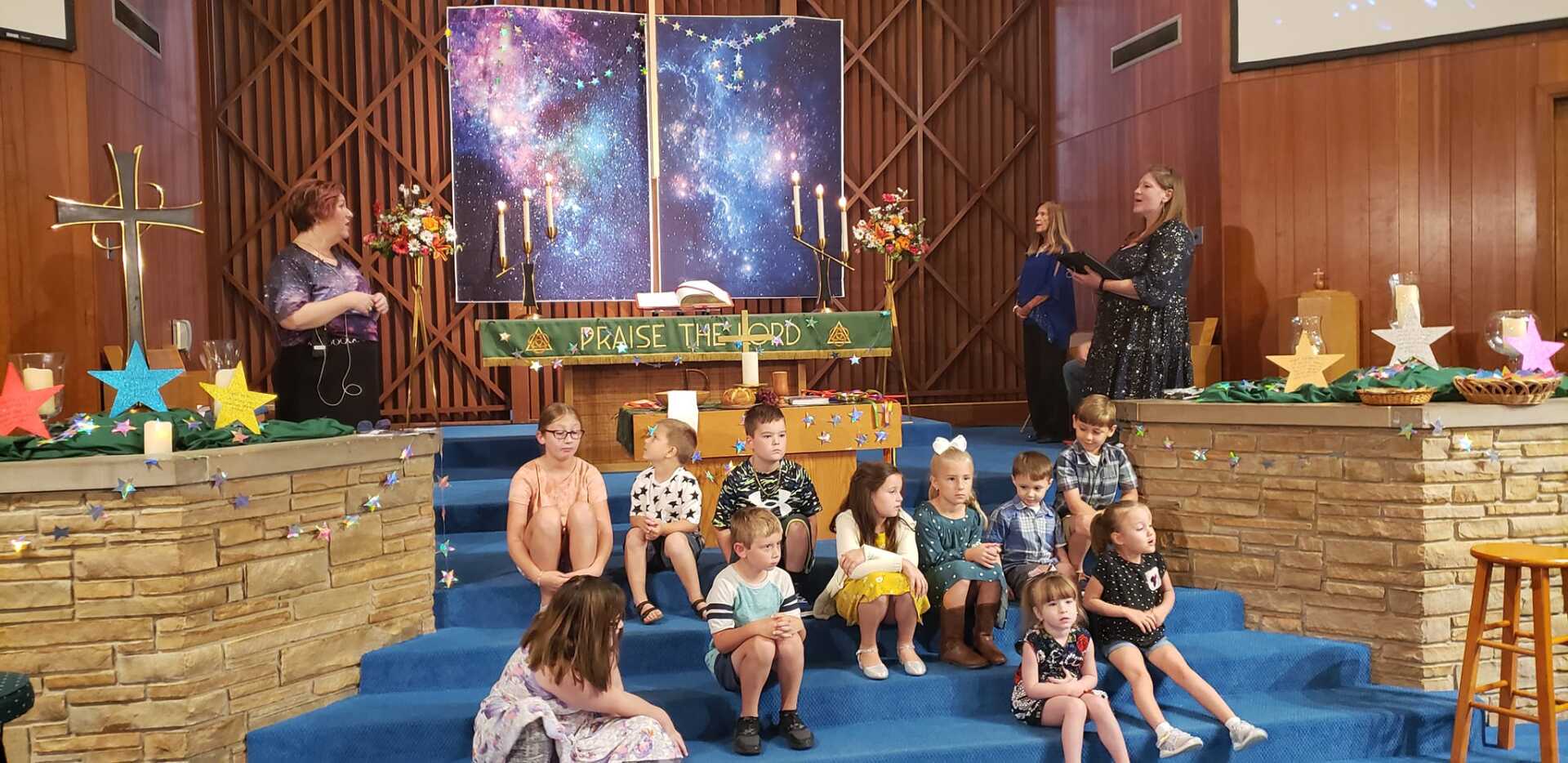 Children and adults in a church; kids seated on blue steps, others standing near a decorated altar, with a starry backdrop.