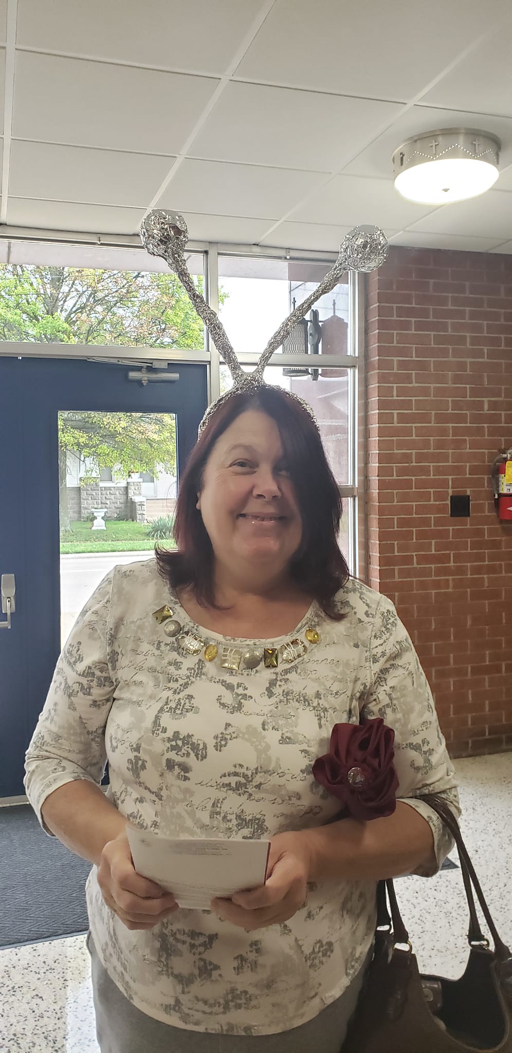 Woman wearing antennae headpiece smiles. She holds a paper, wears a floral top, stands near a brick wall and doorway.