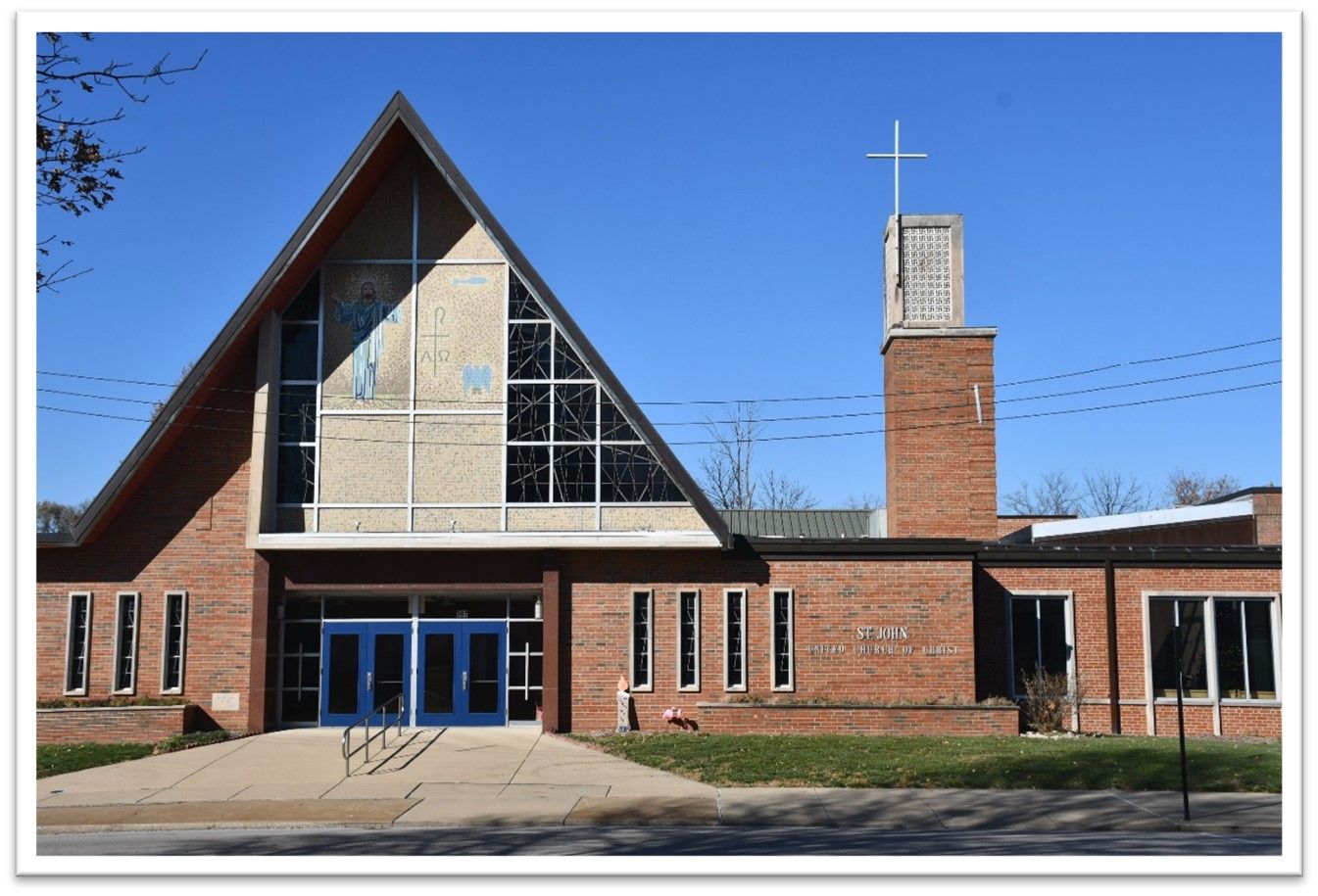 Brick church building with blue doors, triangular roof, and tall steeple topped with a cross.