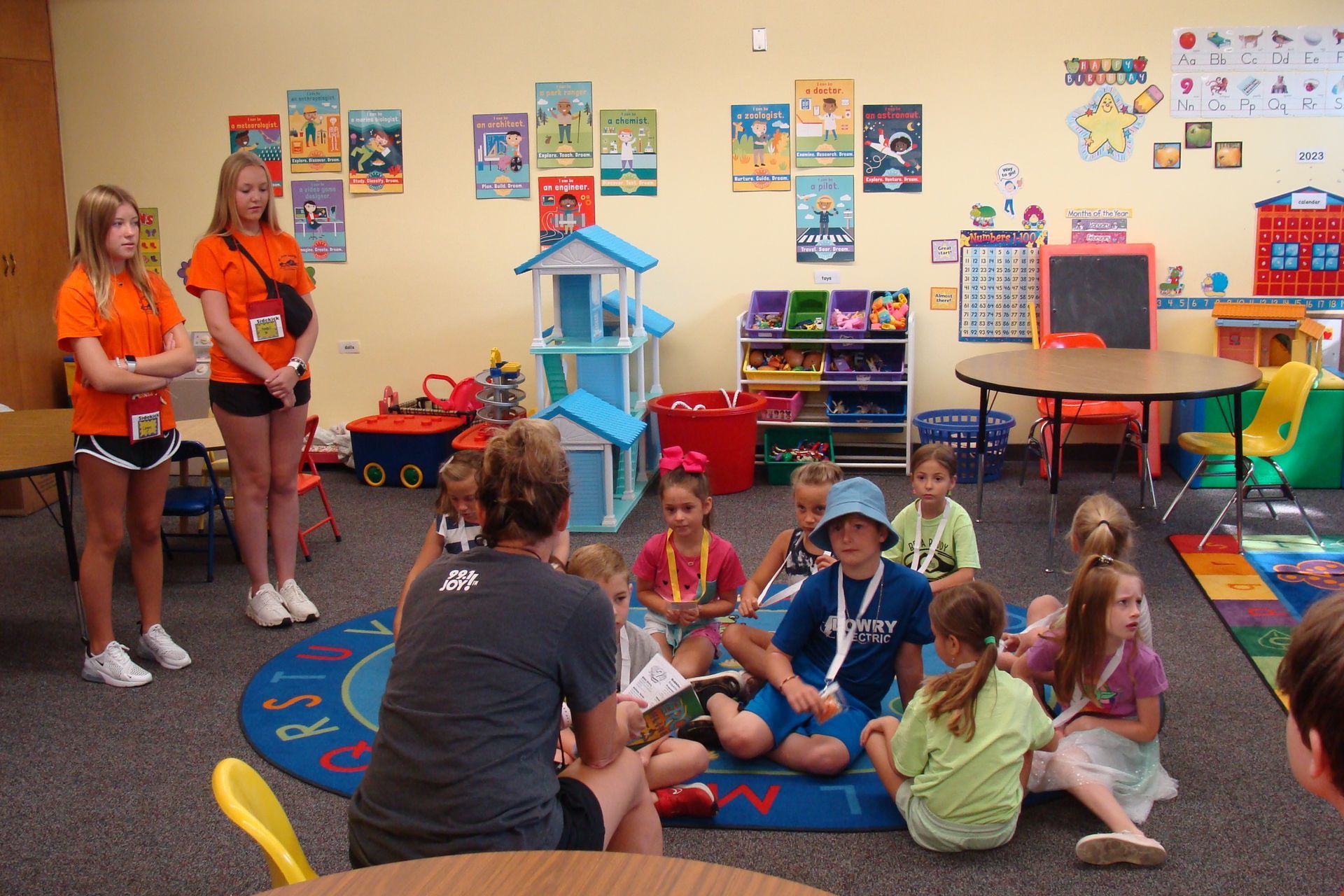 Children and adults in a colorful classroom. One adult reads to kids sitting on a rug; two teens stand nearby.