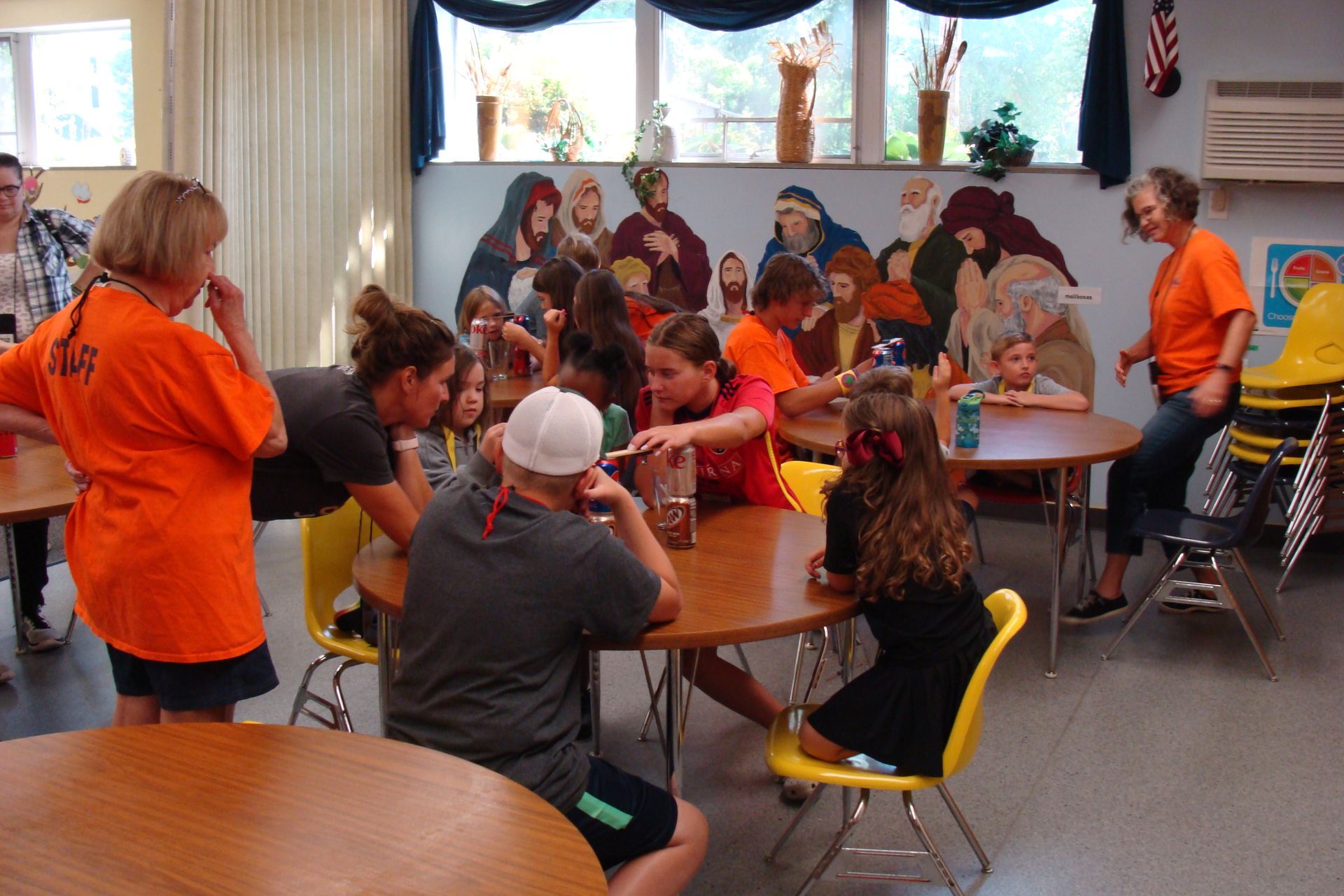 People gather around tables in a room. Some wearing orange shirts. A colorful mural is on the wall.