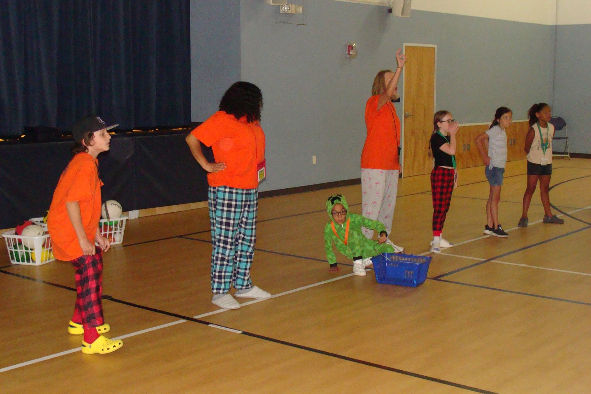 Children in pajamas playing a game in a gym; some wearing orange shirts, others watching.