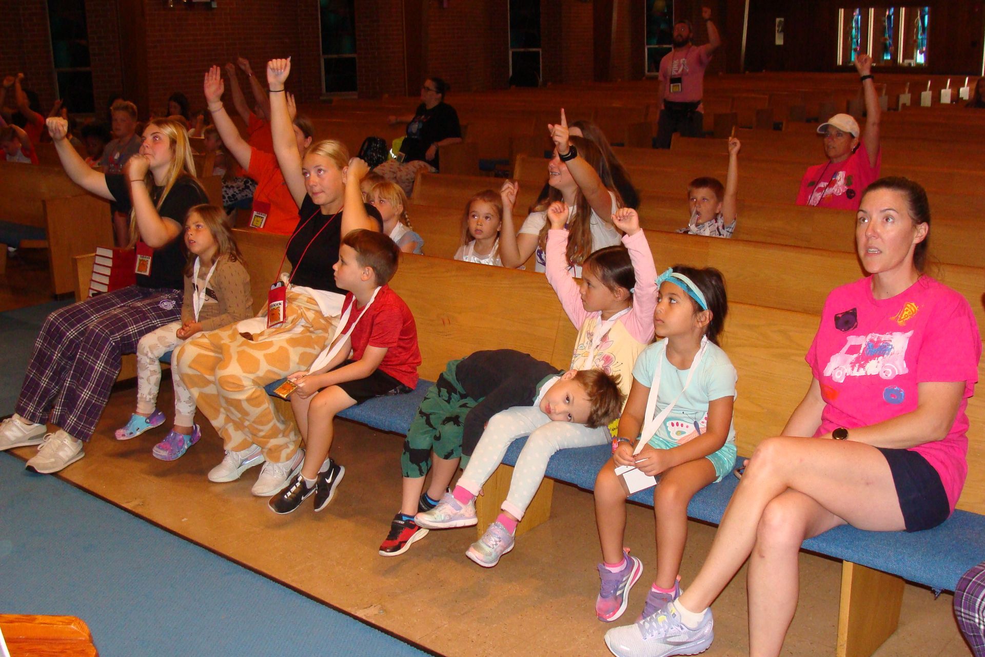 Children and adults raise hands in a church, some sitting, others kneeling. Bright colors, excited expressions.
