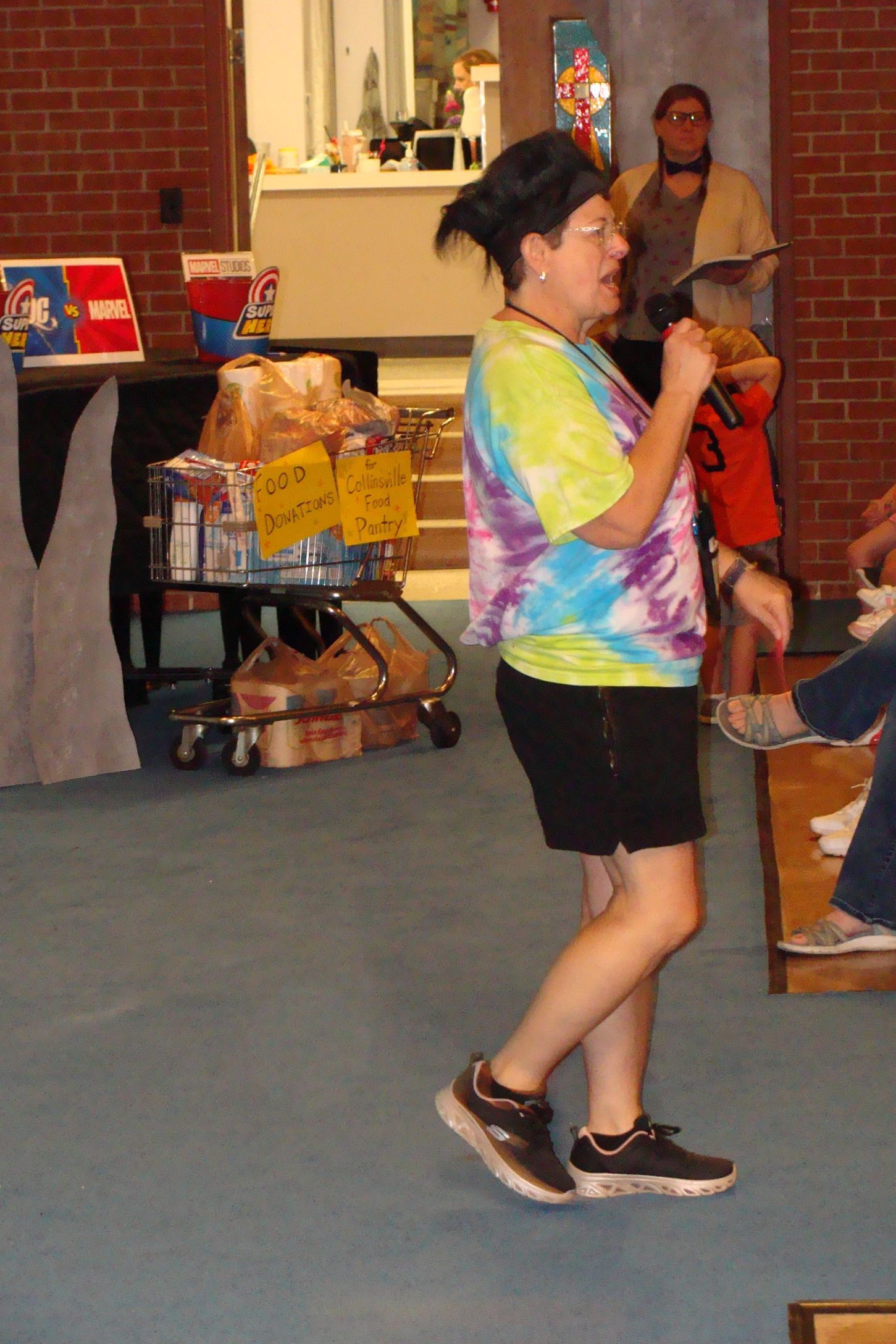 Woman in tie-dye shirt speaking, standing near a cart of food and a seated person.