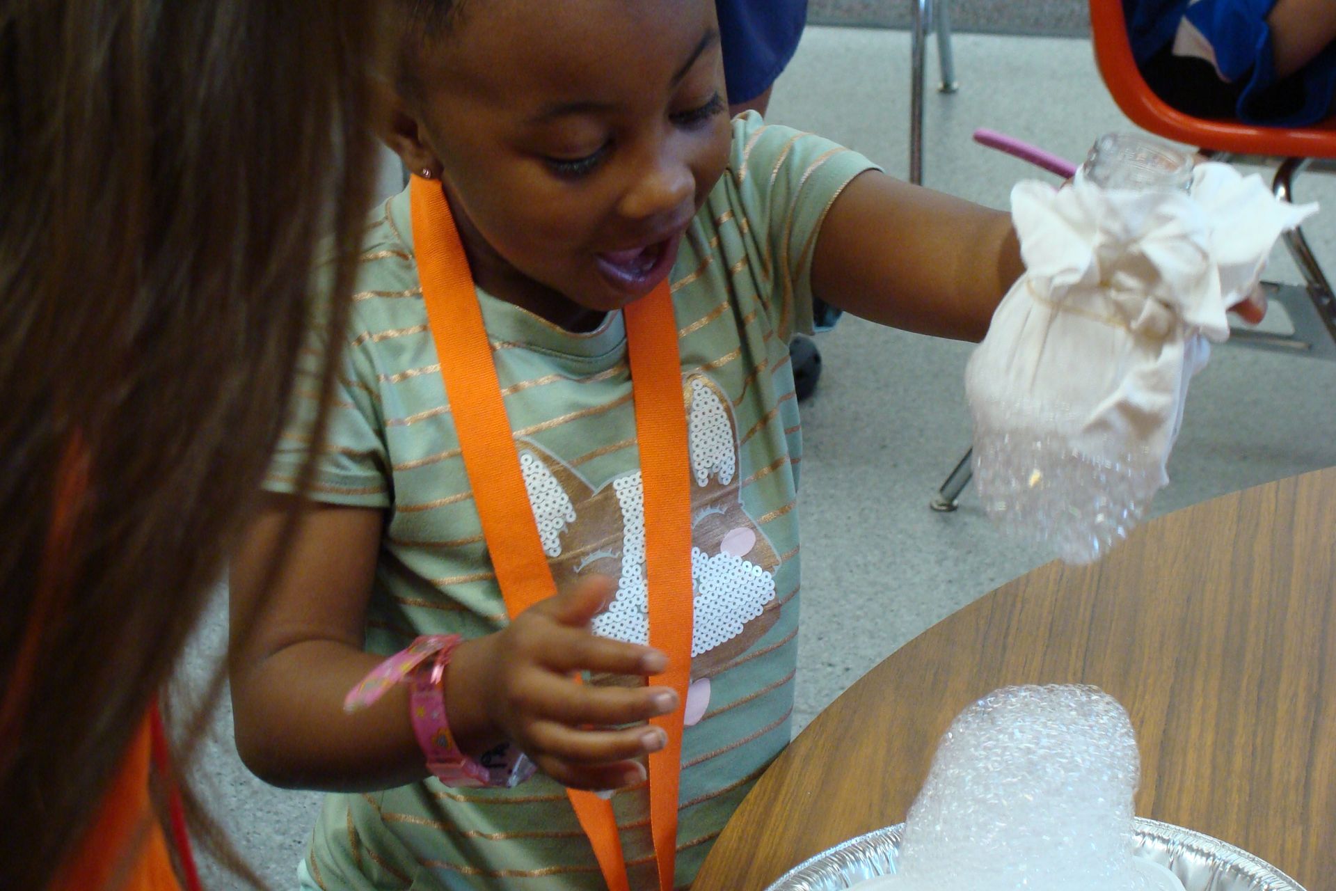 Young girl with an orange lanyard playing with white bubbles on a table, looking excited.