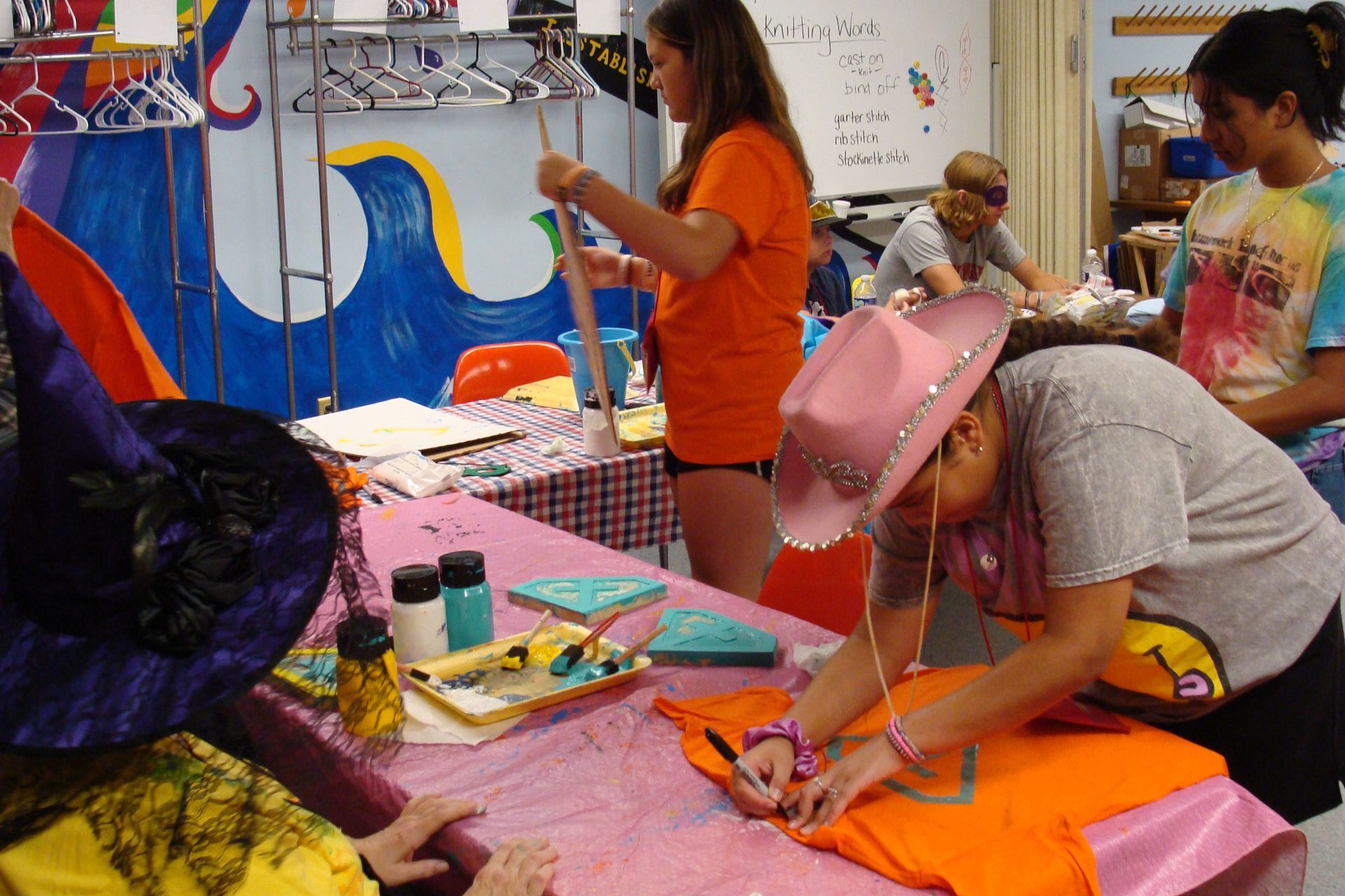People crafting at a table in a workshop, decorating t-shirts, one wearing a pink cowboy hat.
