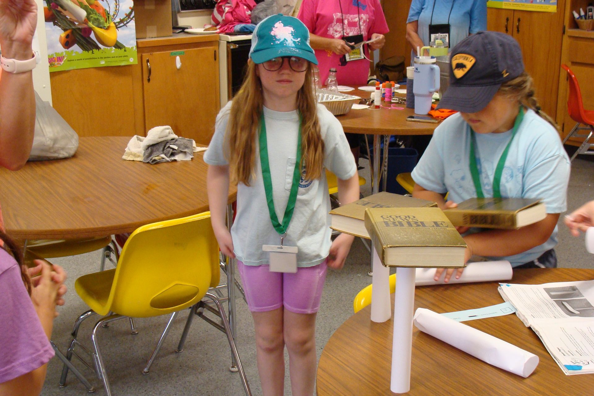 Two girls building with books and tubes in a classroom. One wears a hard hat, the other a baseball cap.