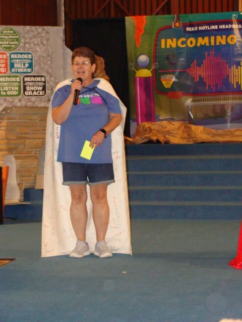 Woman in cape speaking at event. Stage backdrop, signs, blue flooring, cone.