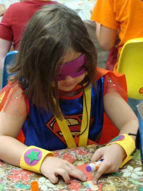 Girl in superhero costume crafting at a table; wearing a cape, mask, and wristbands.