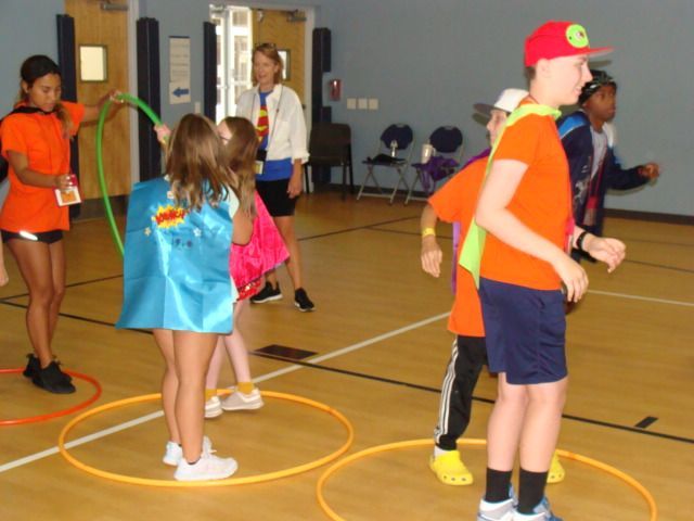 Children in capes playing games with hula hoops in a gym; one adult watches.