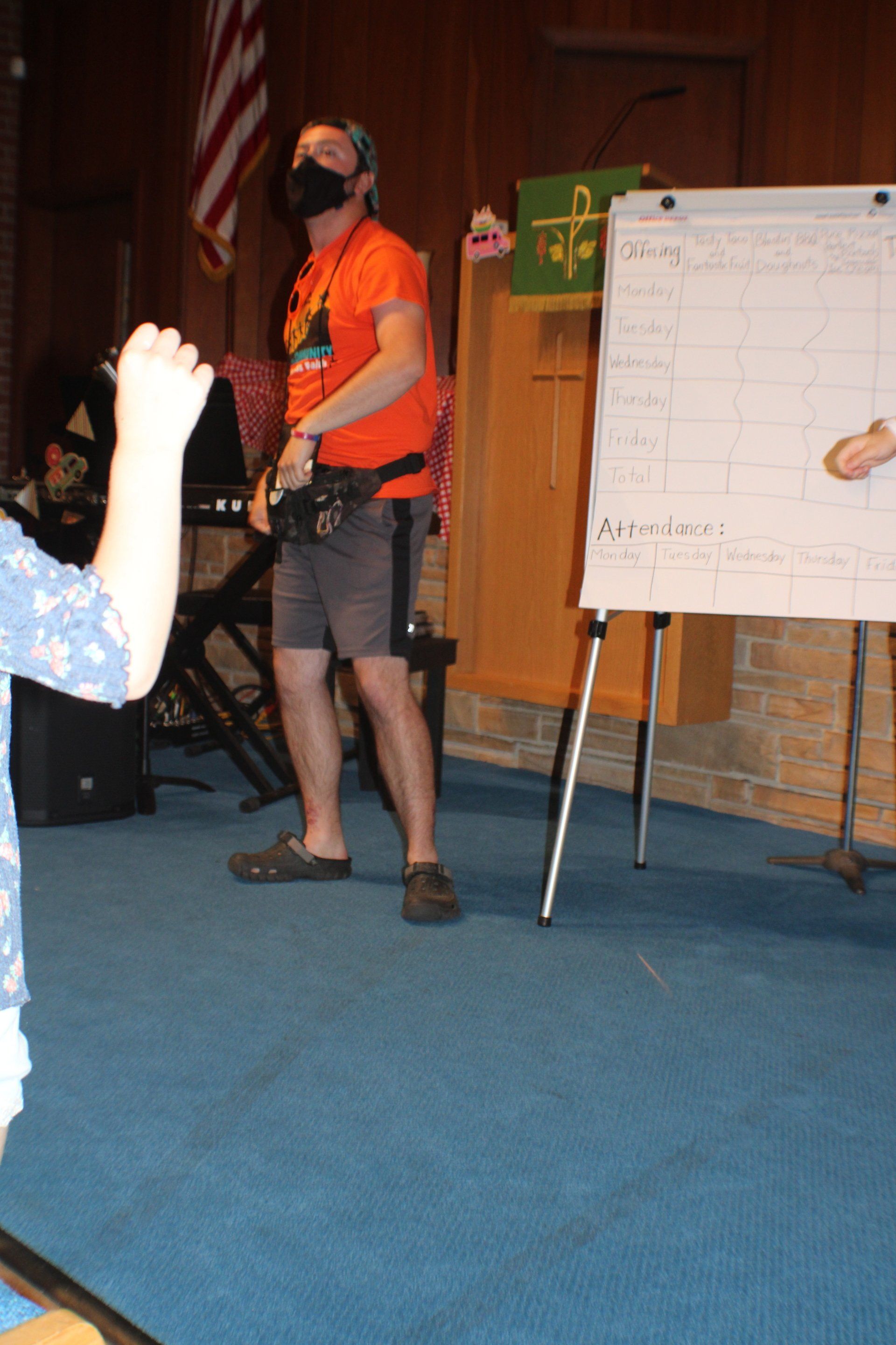 Man in orange shirt and shorts stands in front of a whiteboard, wearing a mask, gesturing.