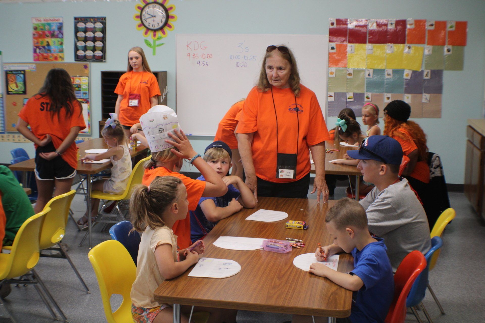 Children and staff in orange shirts at tables, doing crafts in a classroom.