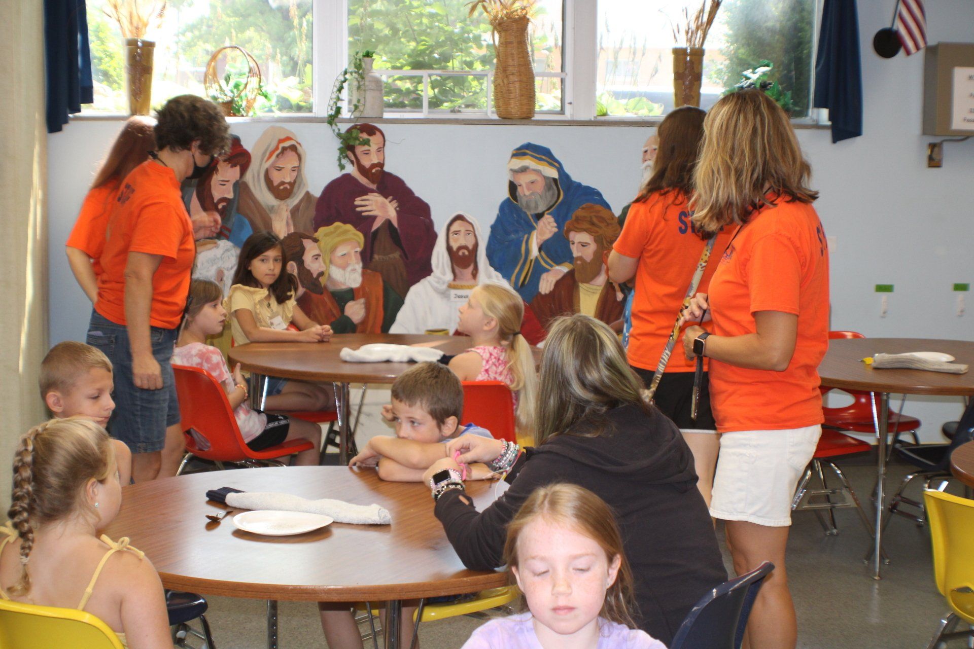 Children and adults gather around tables in a classroom, with cutouts of Jesus and his disciples in the background.
