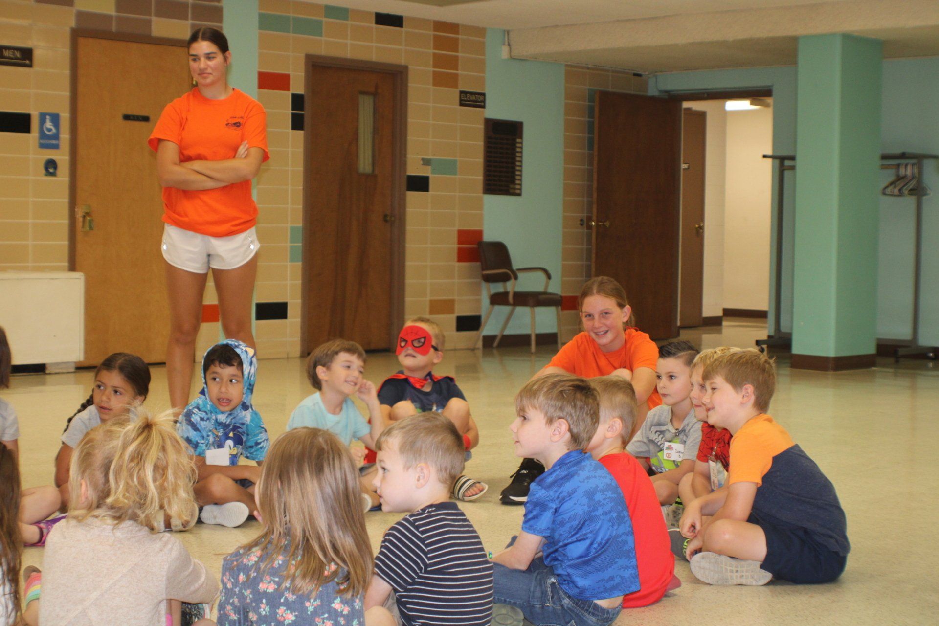 A group of children sitting in a circle listening to two adults, in a room.