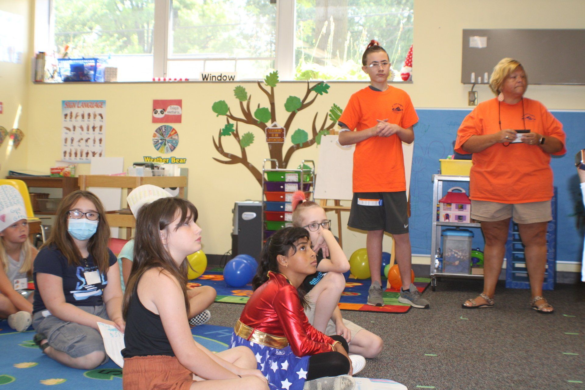 A group of children seated on a carpet, listening to two adults in orange shirts.