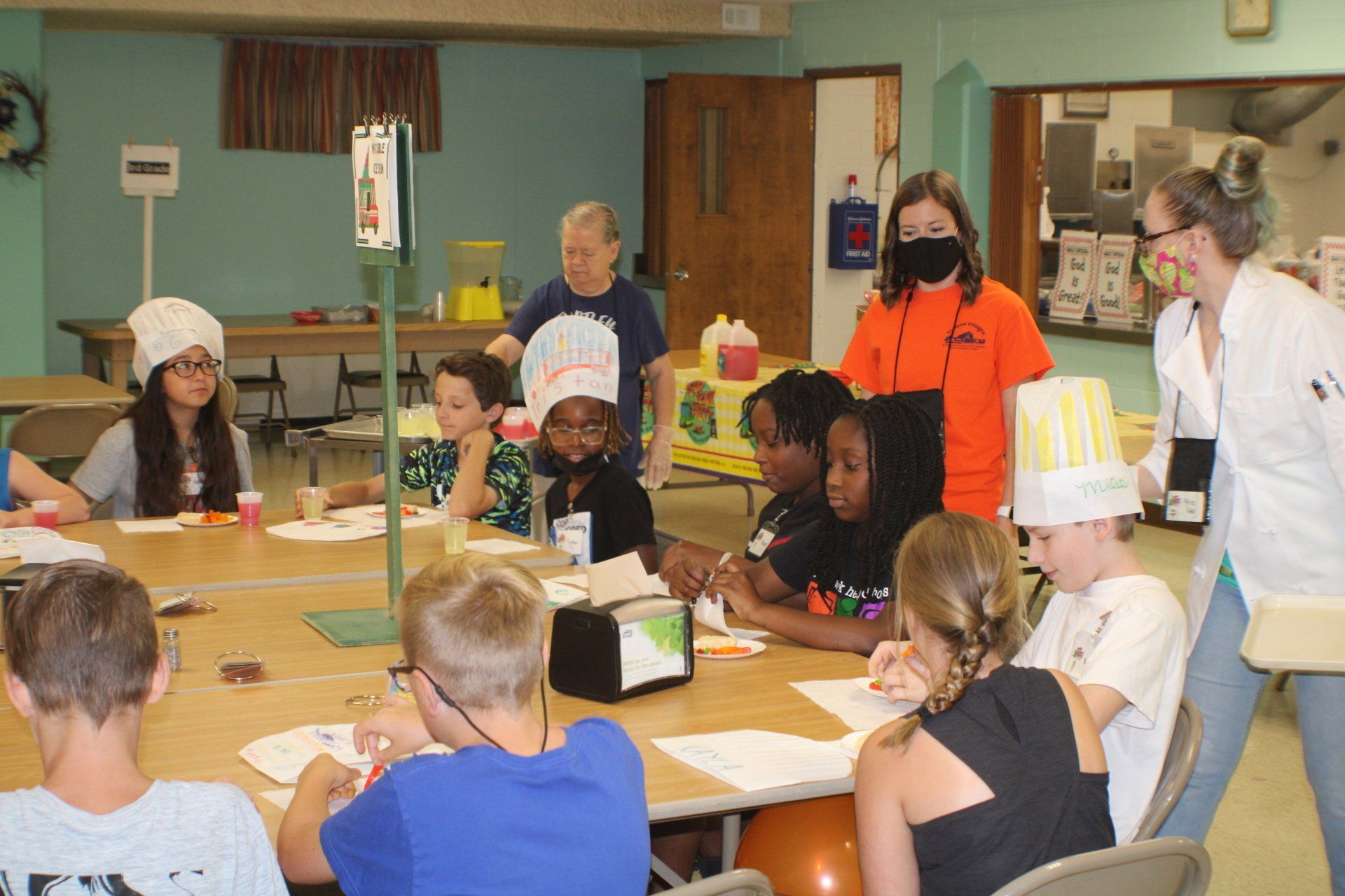 Children and adults in a cooking class, wearing chef hats, seated at a table in a kitchen.
