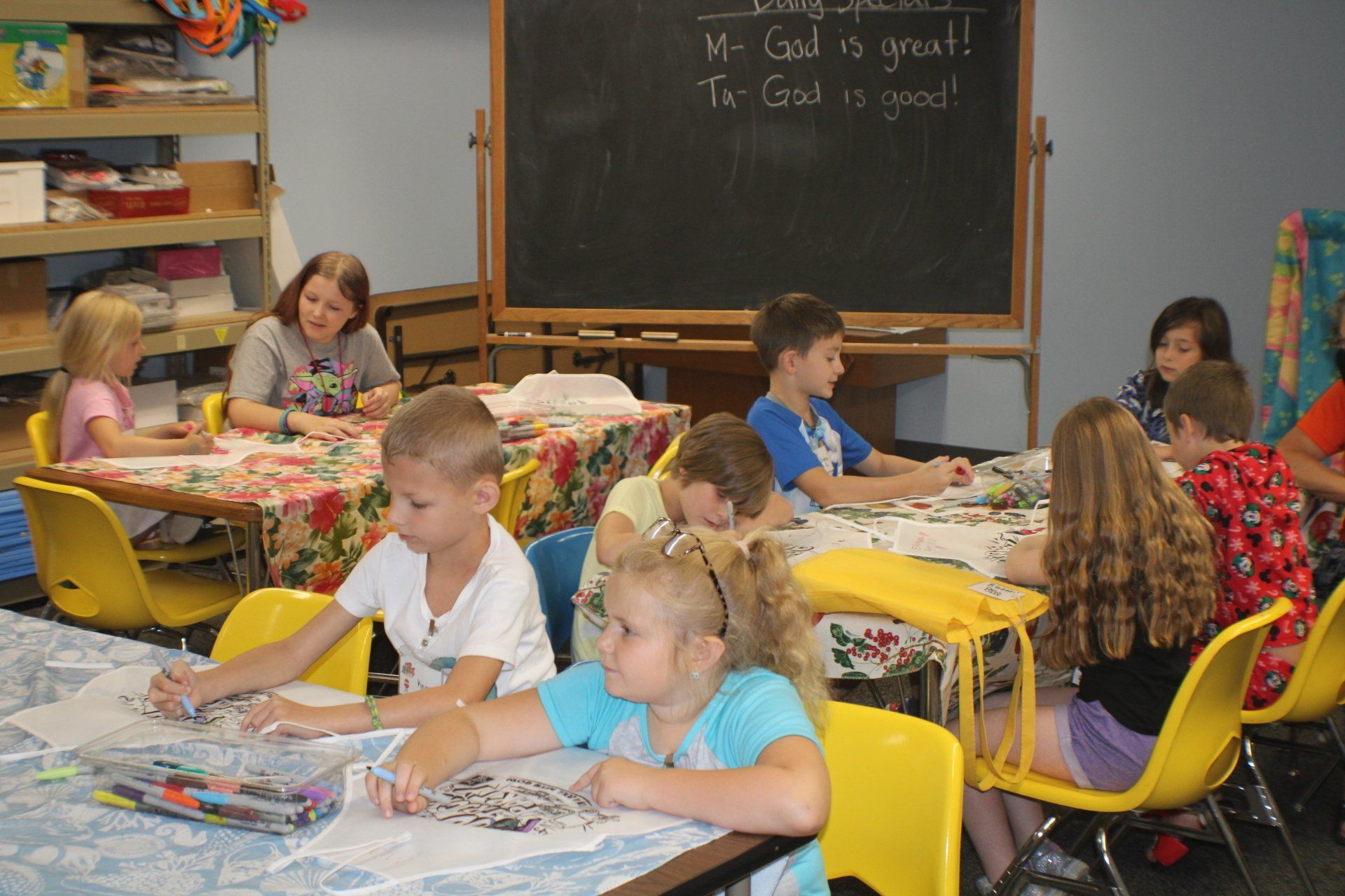 Children coloring at tables in a classroom; a teacher supervises.