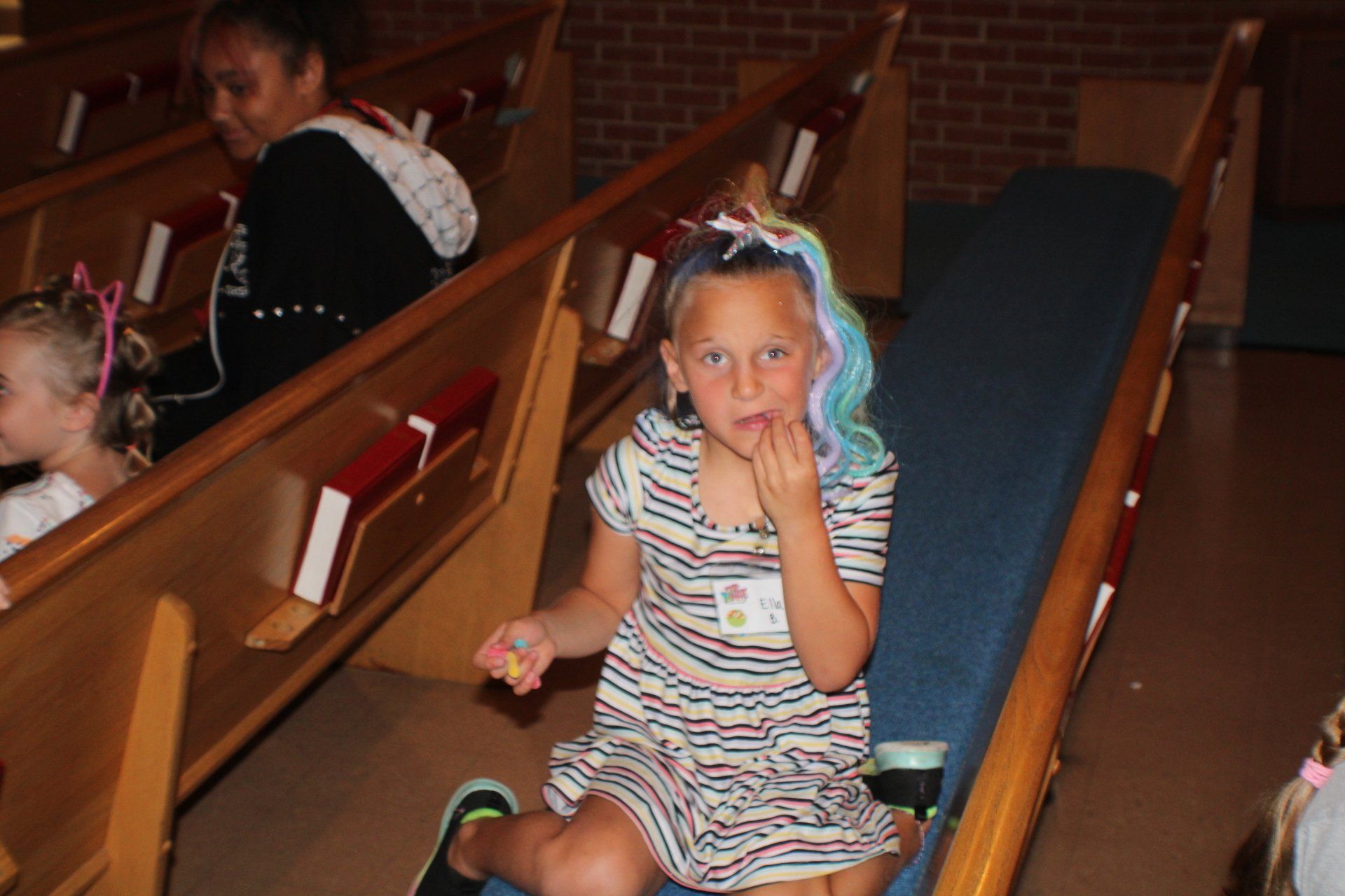 Girl with rainbow hair eating in a church pew, looking at the camera.