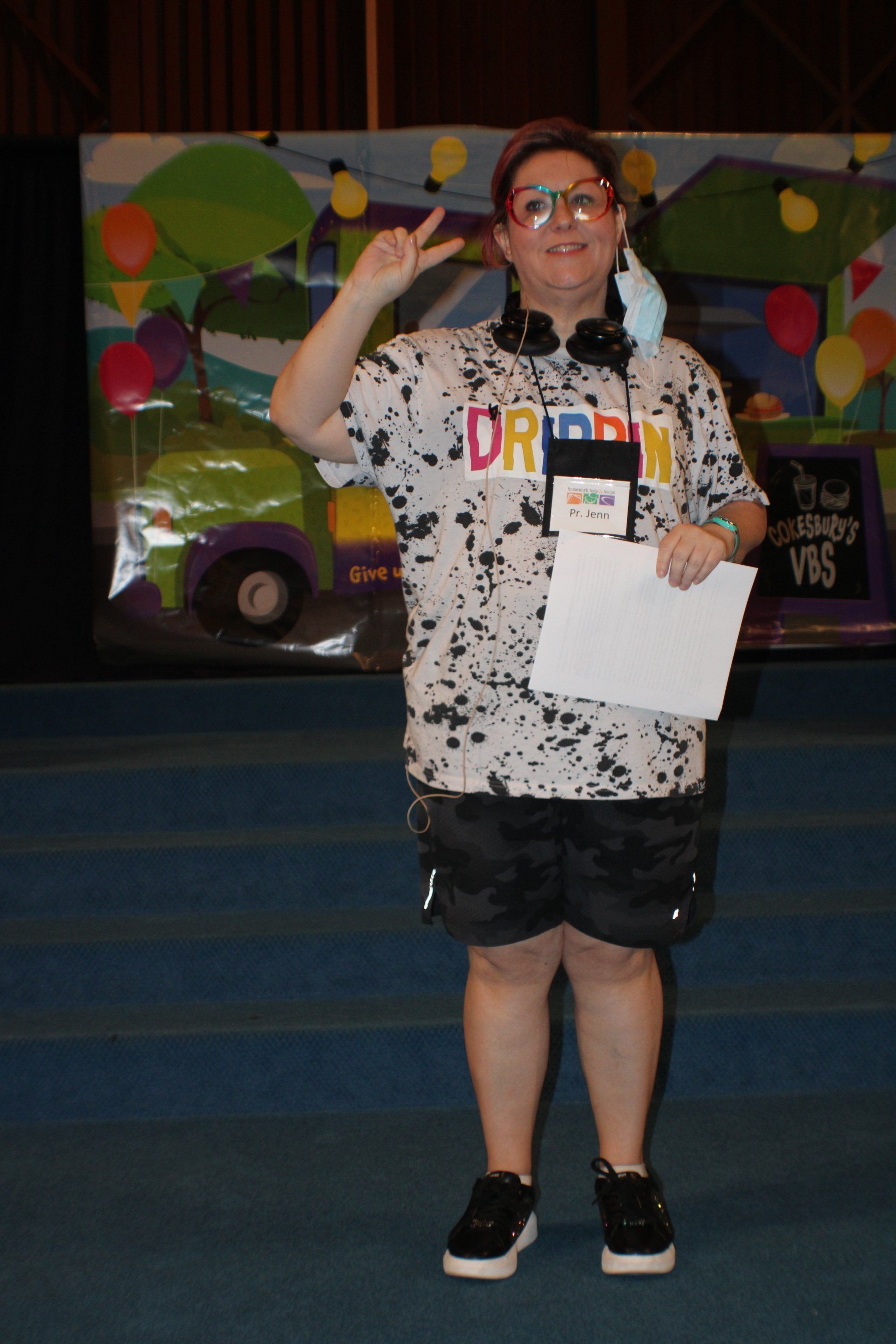 Person in a speckled shirt with headphones, making a peace sign, holding paper. They stand on stage with decorations.