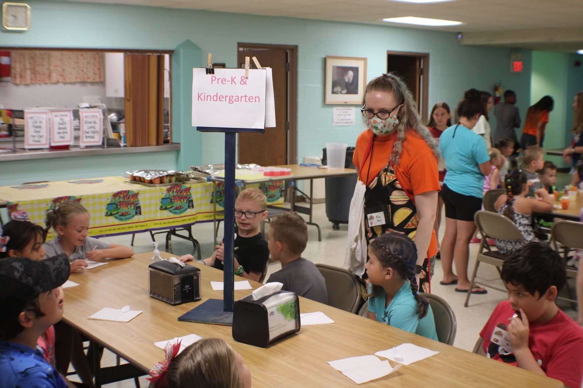 Kindergarten students at lunch with a teacher, sign reads 