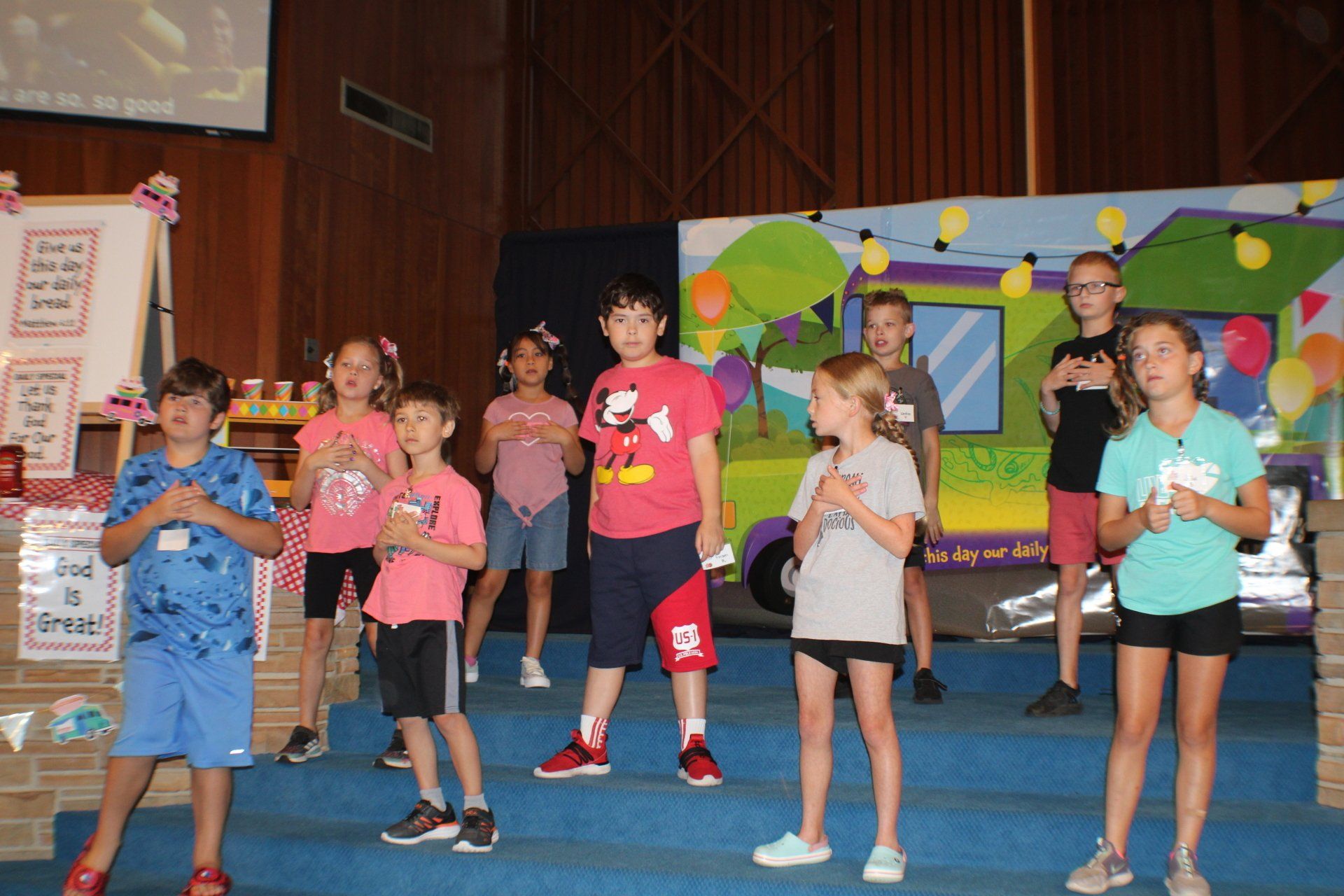Children on a stage singing with hands on chests; a food truck backdrop.