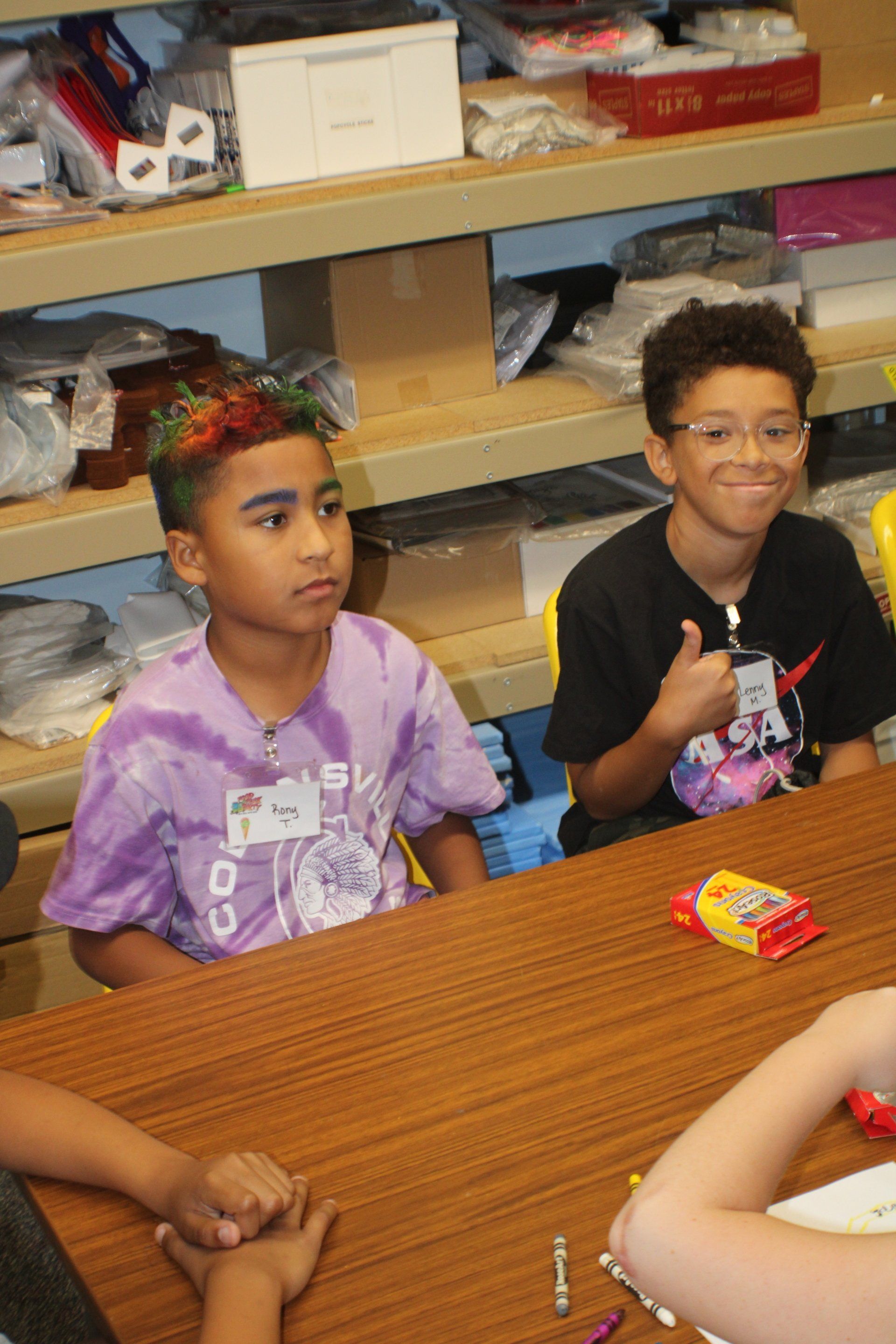 Two boys at a table, one with rainbow hair, the other giving a thumbs-up. Shelves in background.