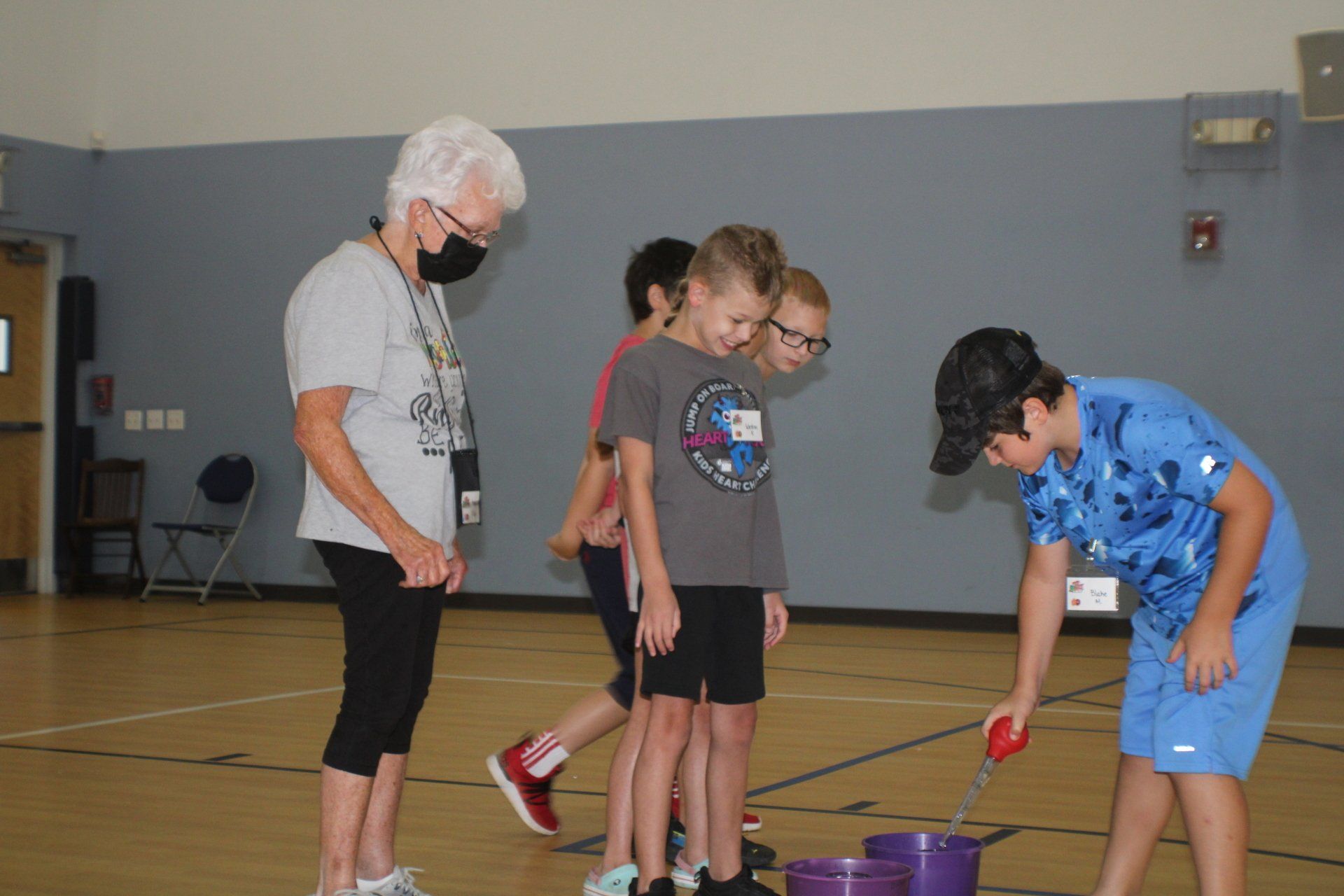 Kids playing a game with a woman in a gym. Boy scoops water from bucket with ladle.