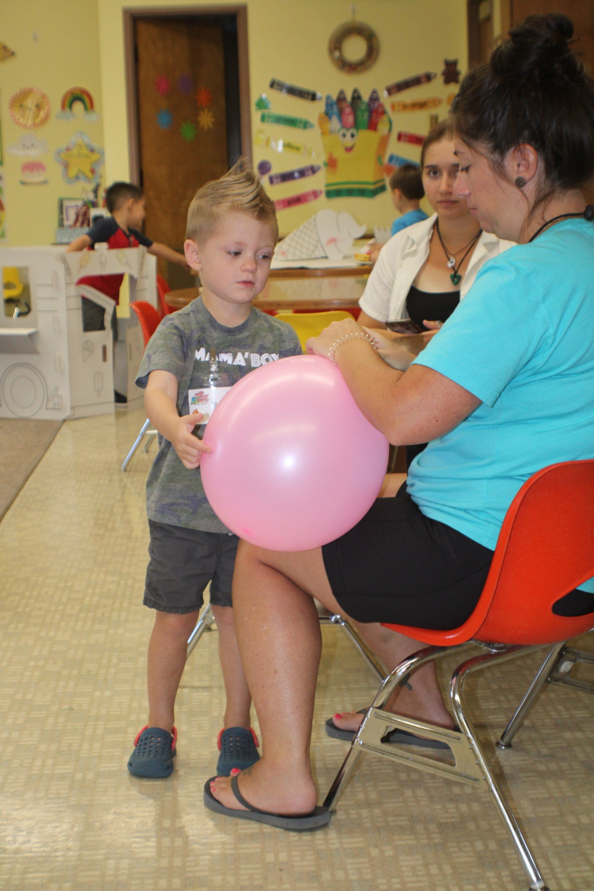 Boy with a mohawk holds a pink balloon. A woman in a blue shirt is holding it. Indoors.
