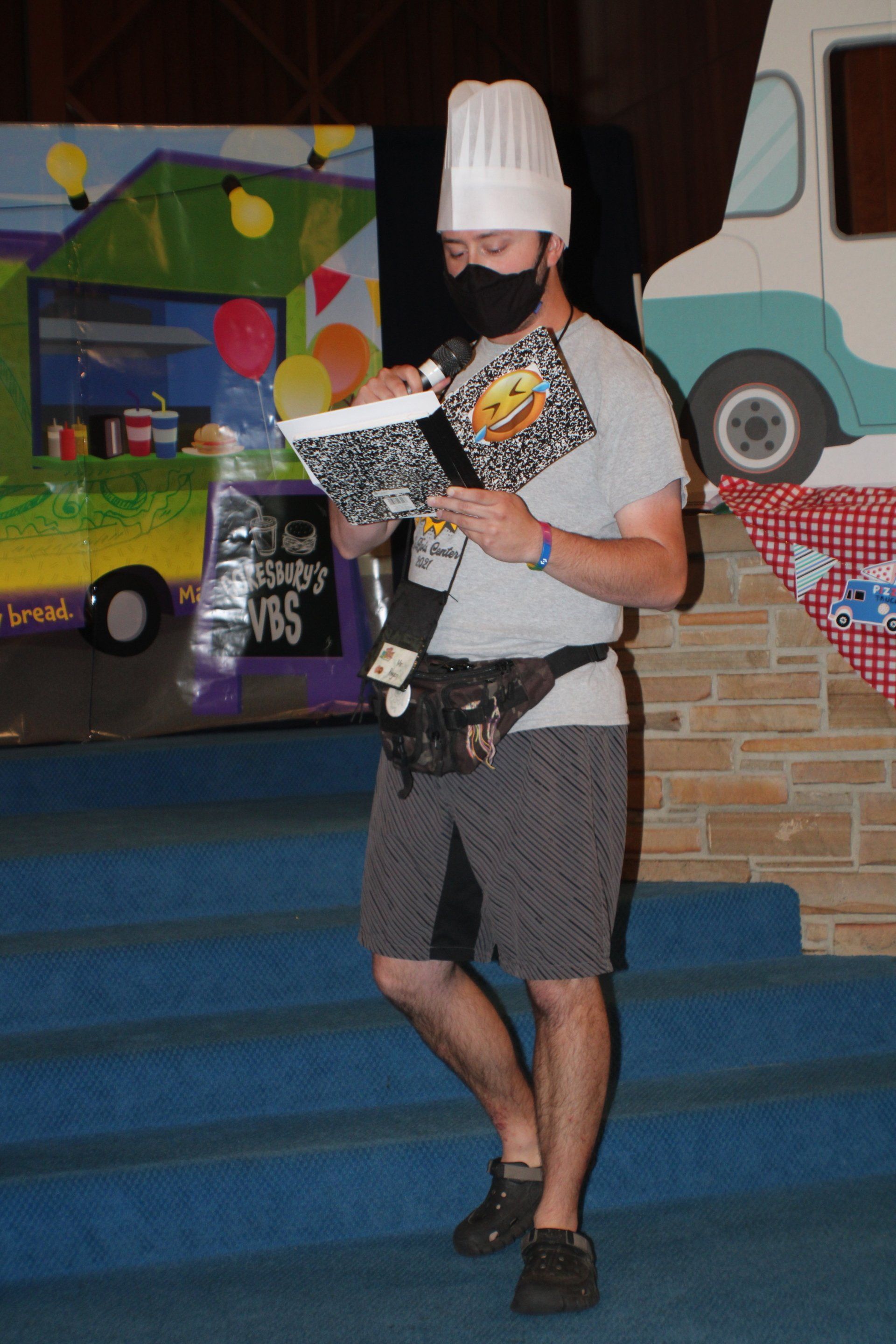 Man in chef hat and mask reading a book, standing on blue stairs, food truck backdrop.