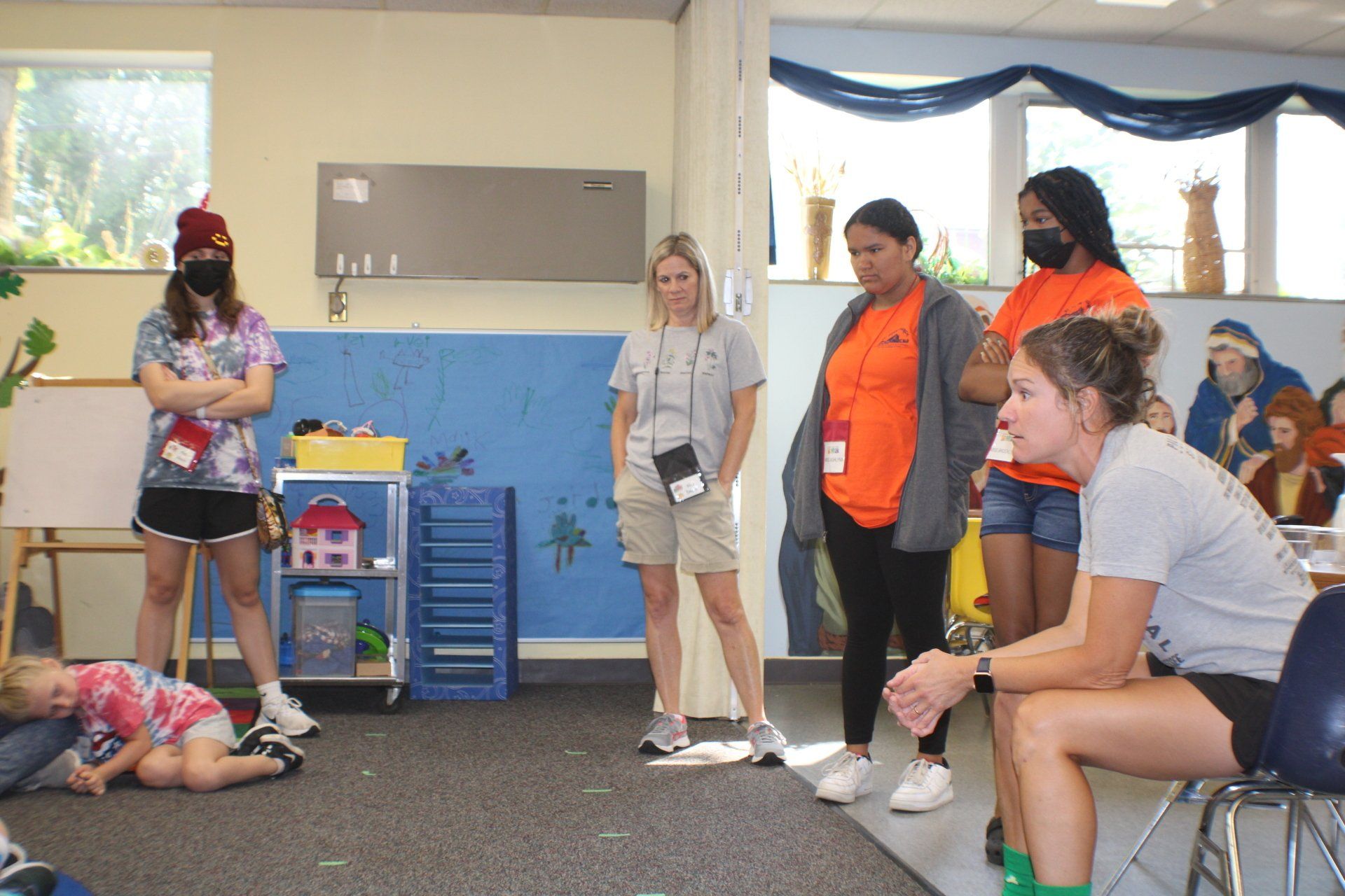 A group of adults in a classroom setting, some masked, with a child on the floor; all appearing attentive.