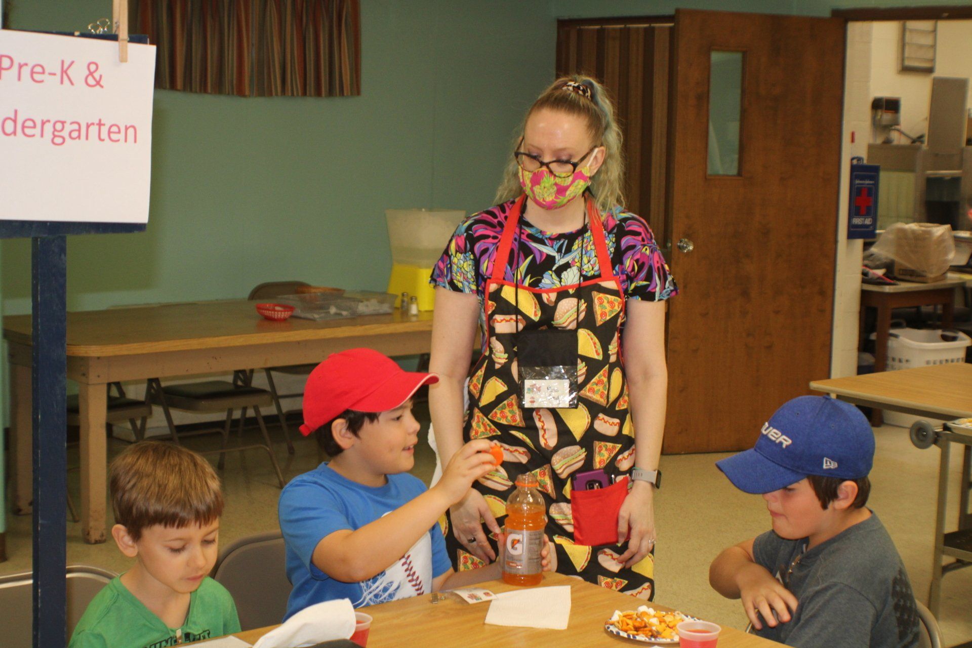 Teacher with mask watches three kids at table; one holds snack, other two wear caps.