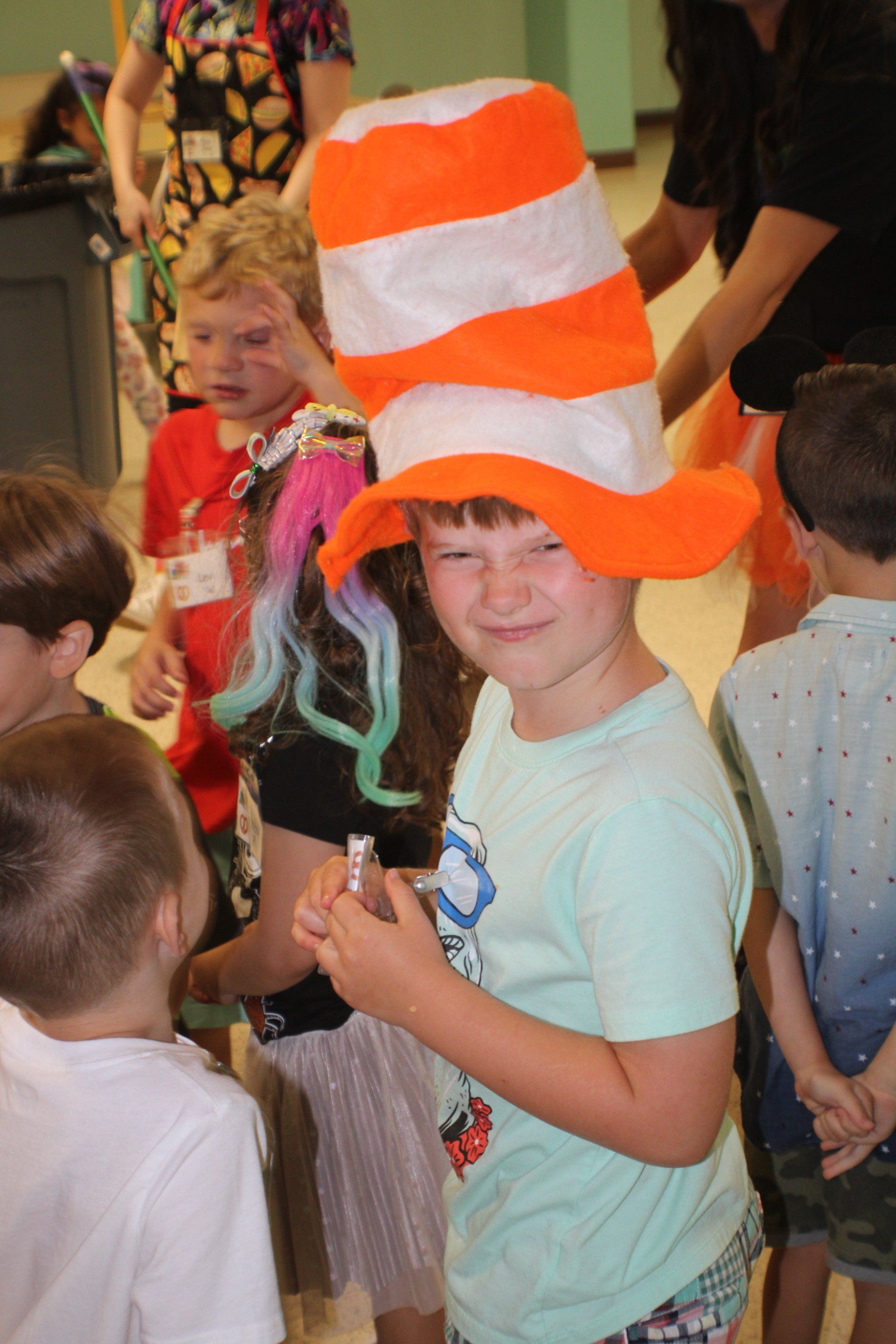Child wearing a tall orange and white hat, smiling, surrounded by other children, possibly a costume party.