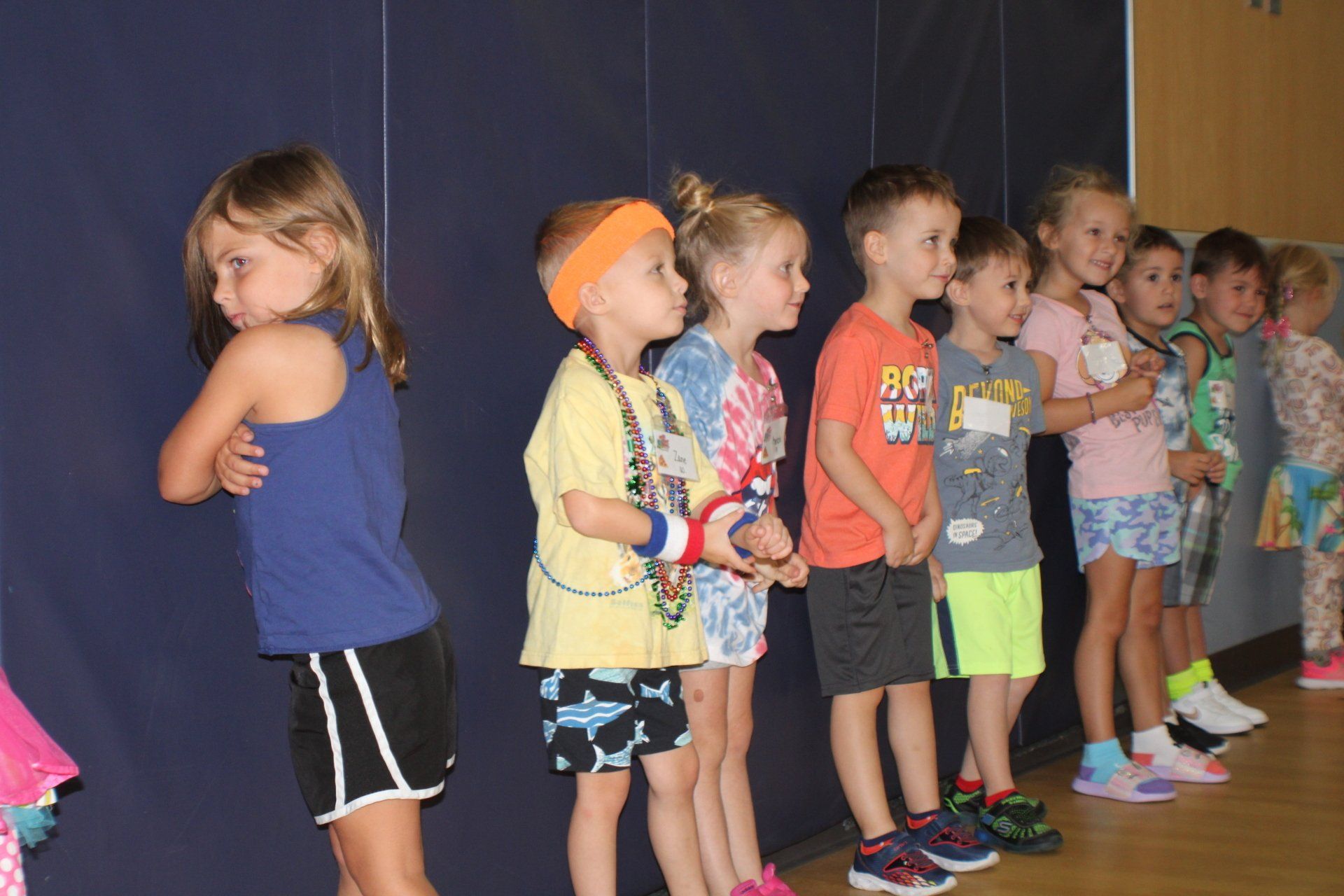 Children in colorful outfits line up against a blue wall, one girl with arms crossed, other kids looking forward.