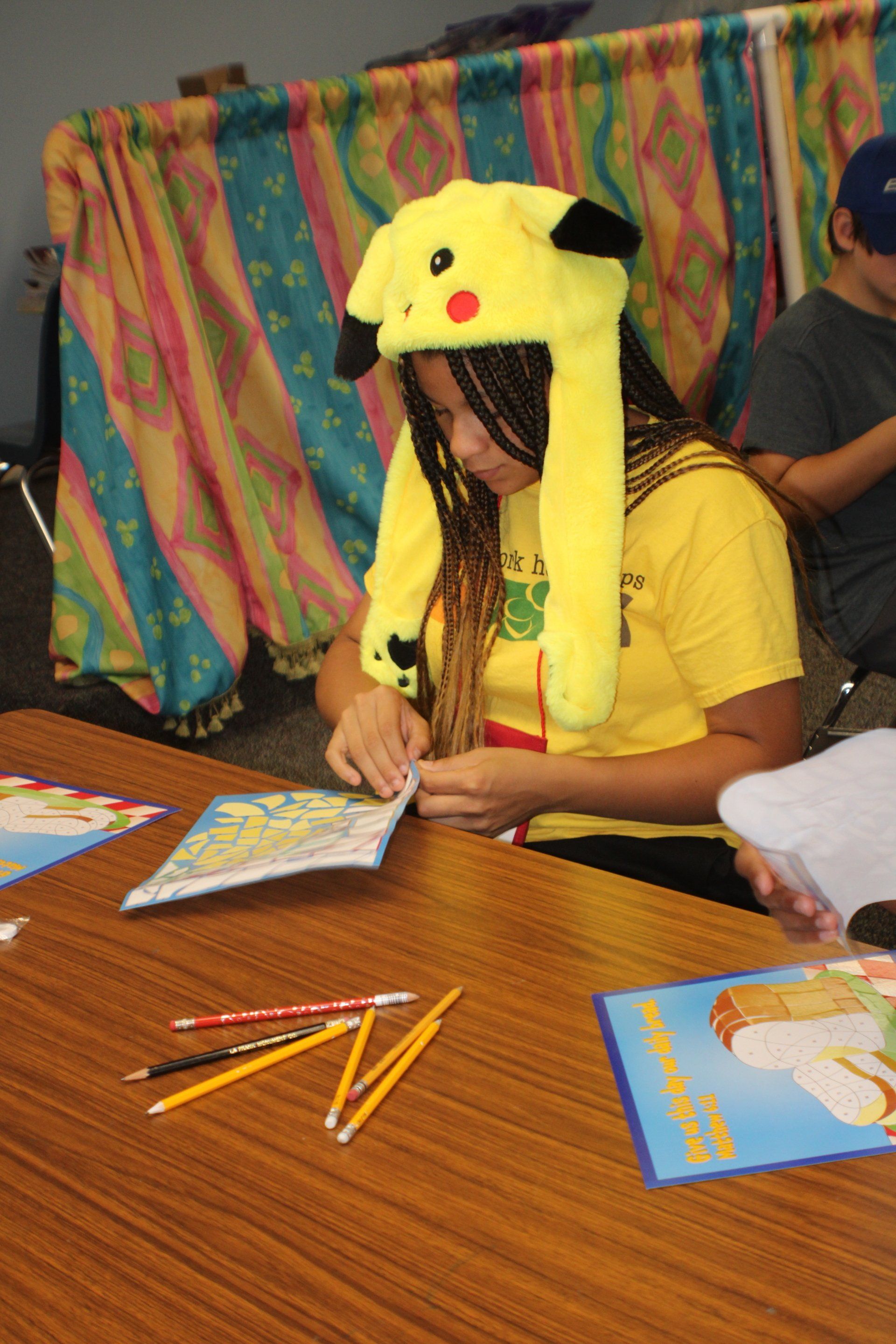 Person wearing Pikachu hat, in yellow shirt, looking at book at a table.