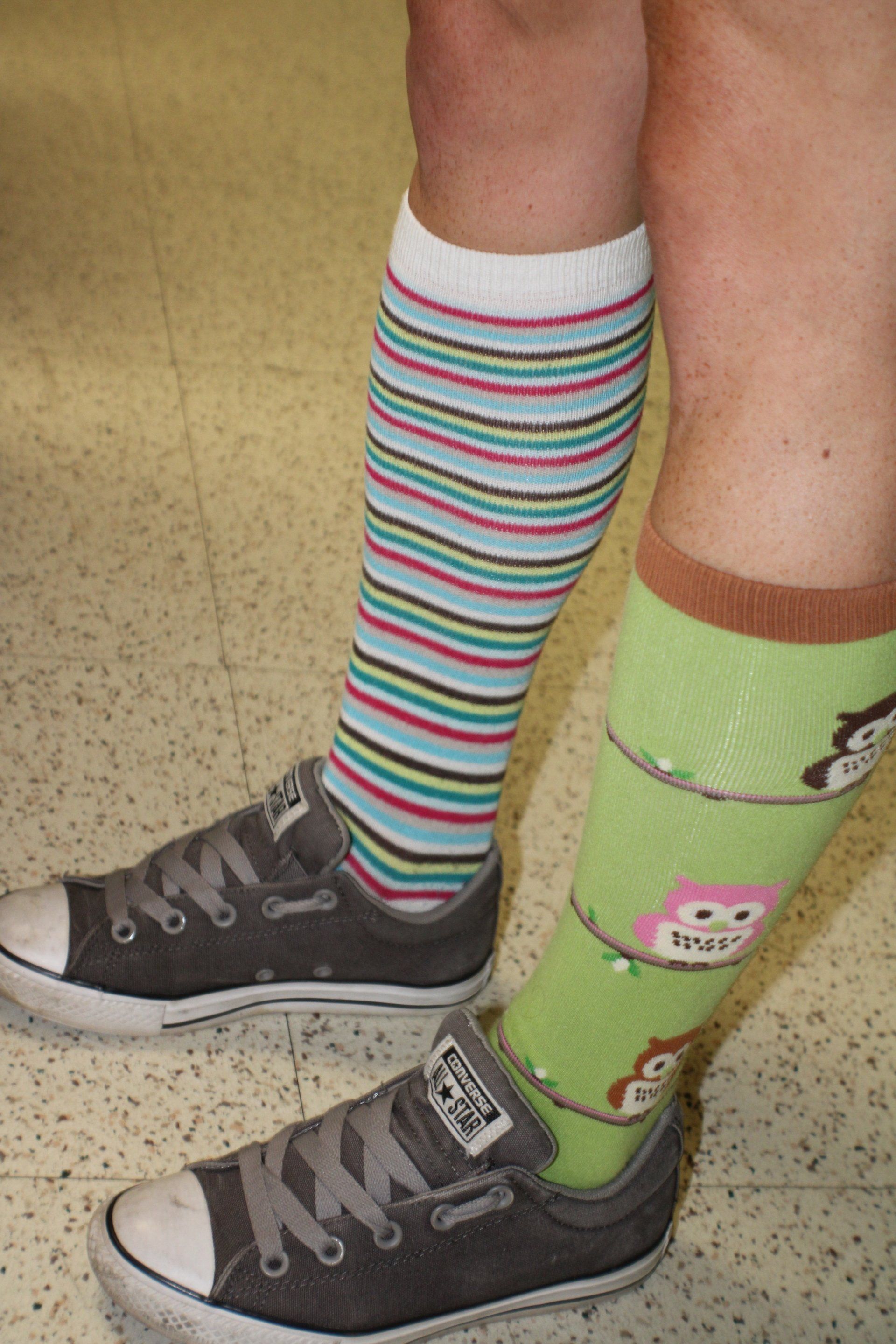 Person wearing mismatched socks (striped and owl-themed) with gray Converse sneakers on a light-colored floor.