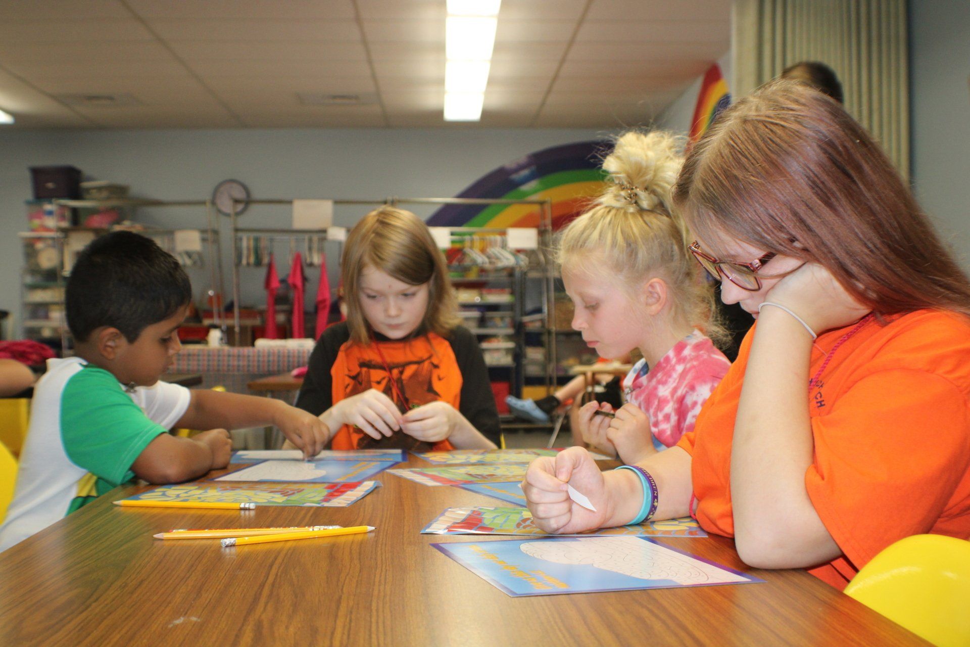 Children and a teen sit at a table, focused on projects. Classroom setting, rainbow mural visible.