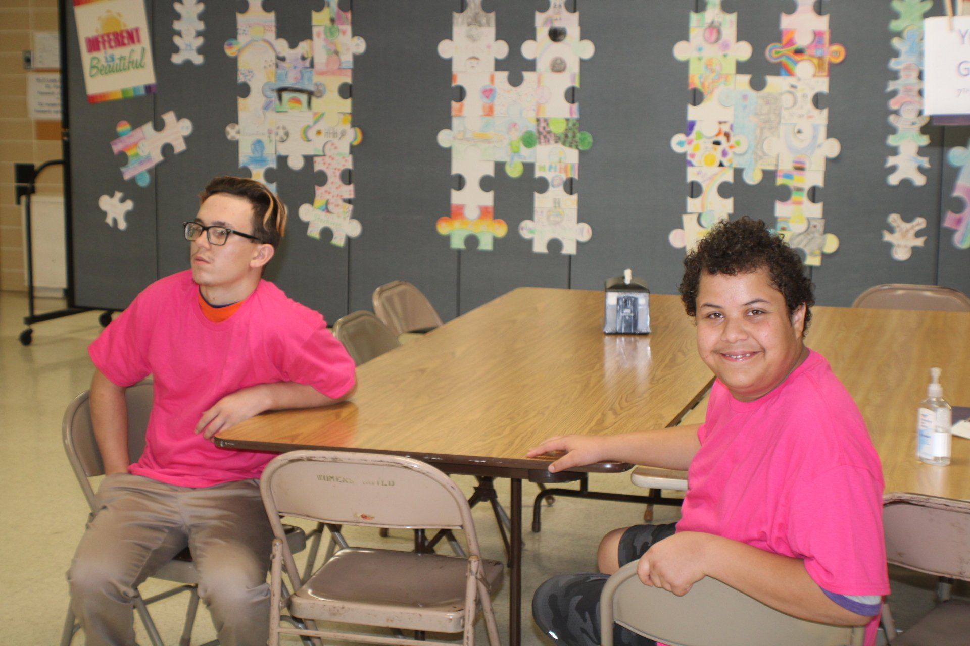 Two people in pink shirts at a table; puzzle art background; one person looks away, the other smiles.