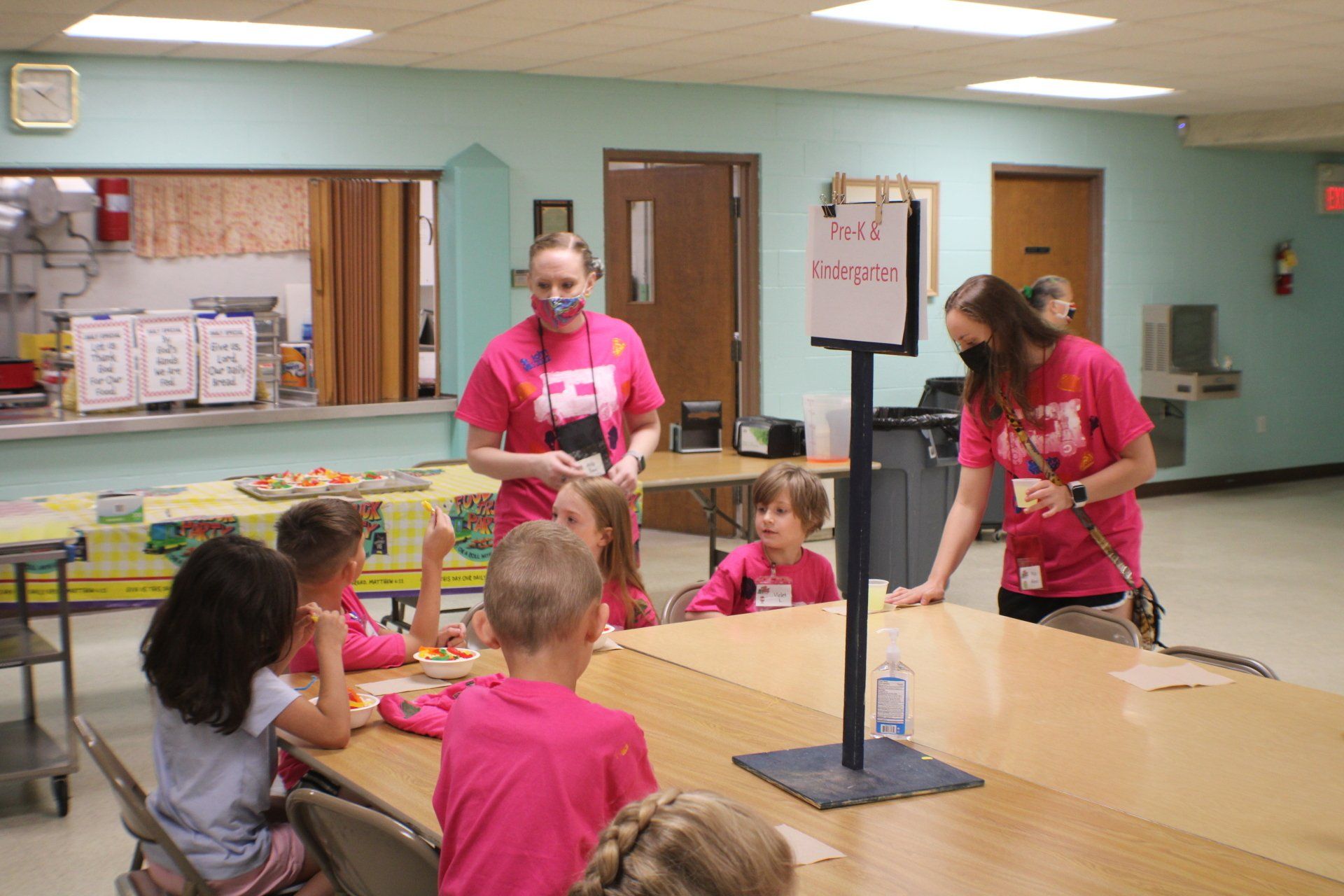 Two women in pink shirts assist children at a lunch table; a sign reads 