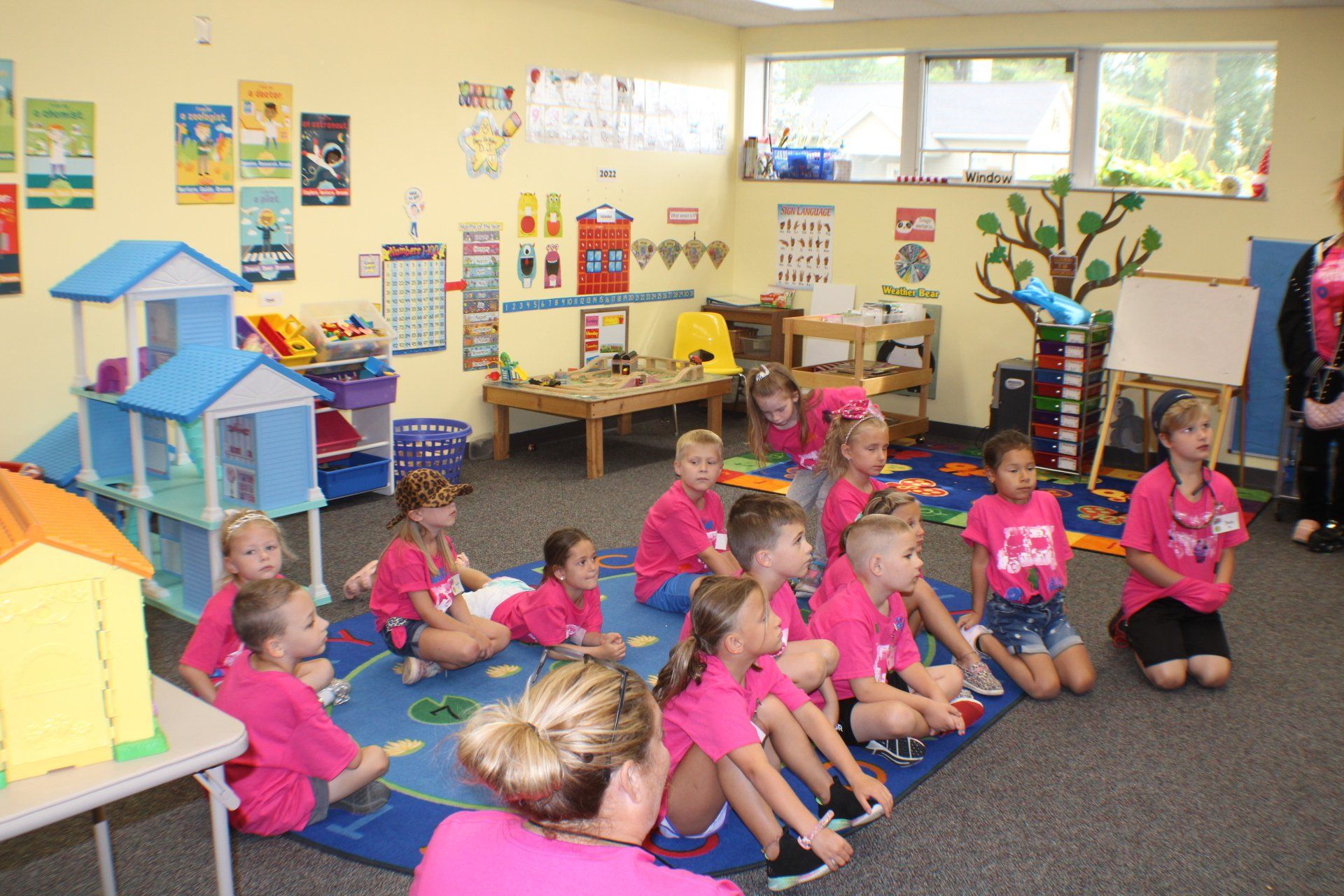 Children in pink shirts sitting on rug in a preschool classroom.