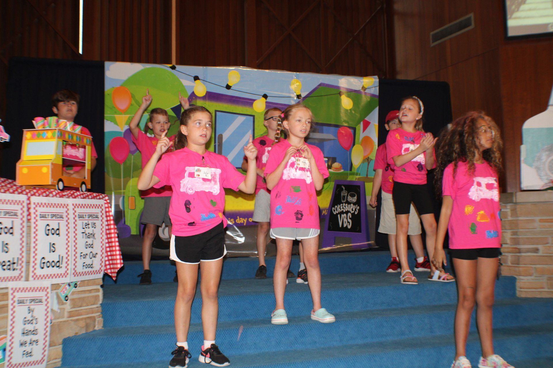 Children on stage in pink shirts with ice cream truck logos, singing. Colorful backdrop, church setting.