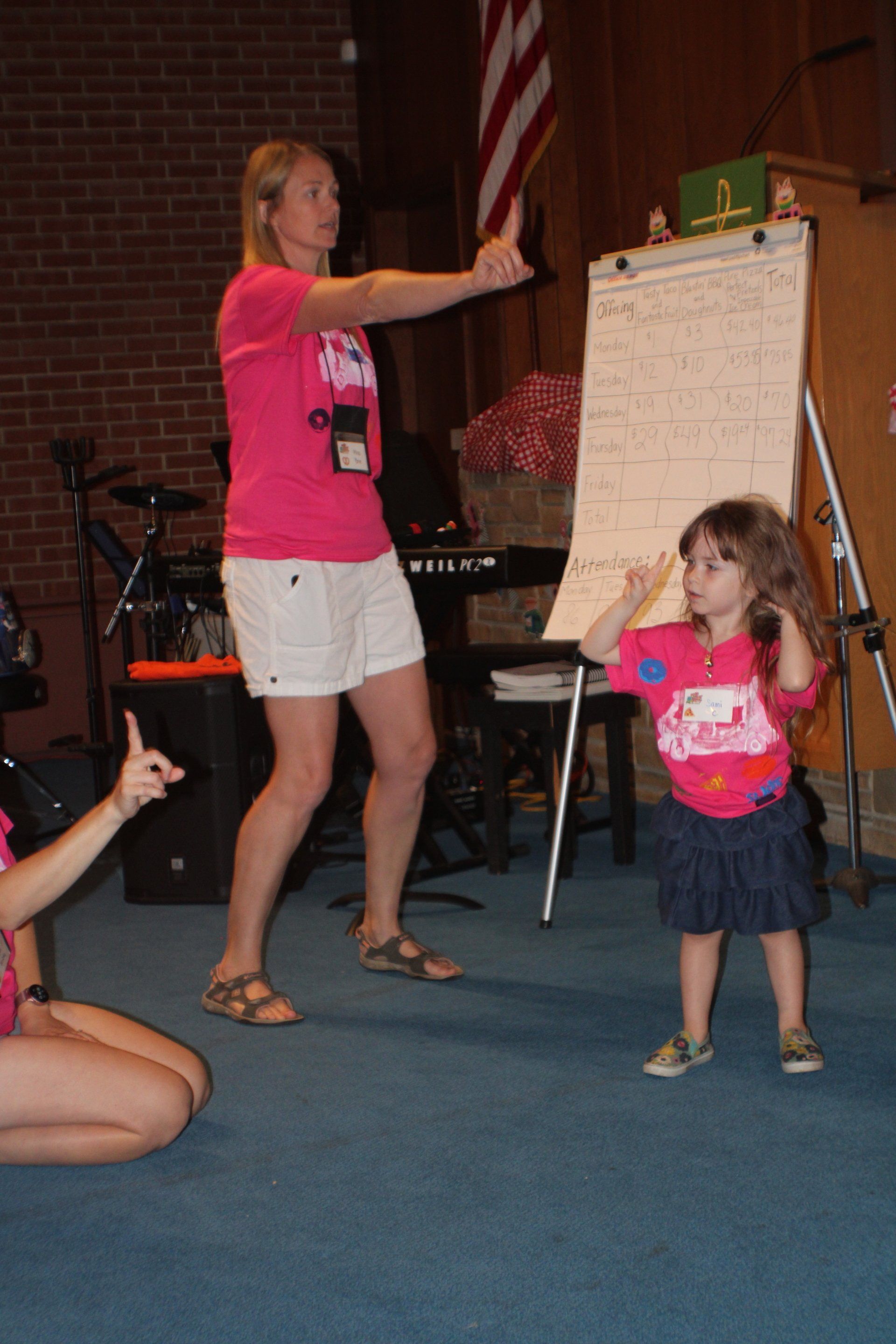 Woman and child in matching pink shirts and white shorts gesturing, in front of a whiteboard and American flag.