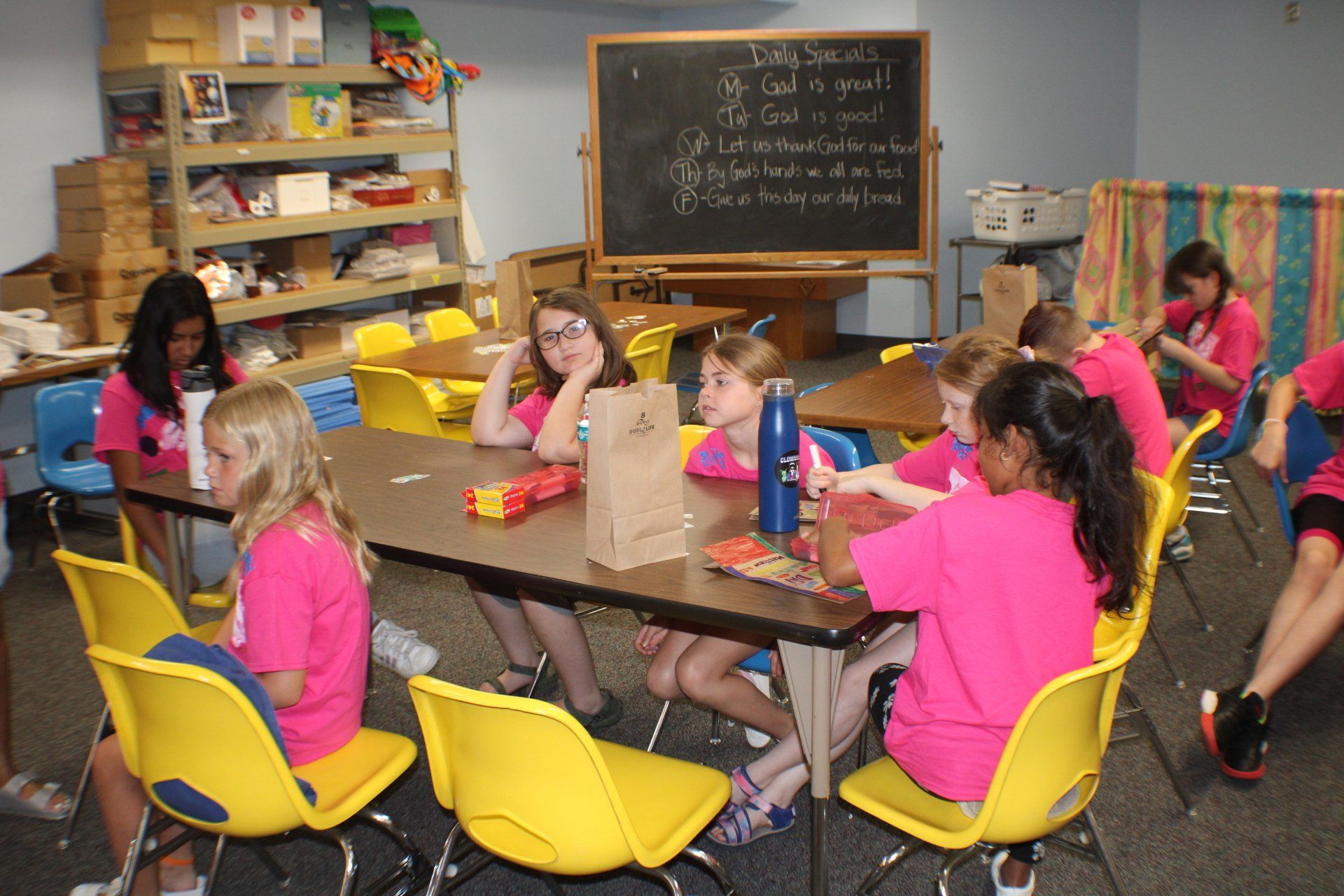 Girls in pink shirts sit at tables, possibly eating, in a classroom with a chalkboard and shelves.