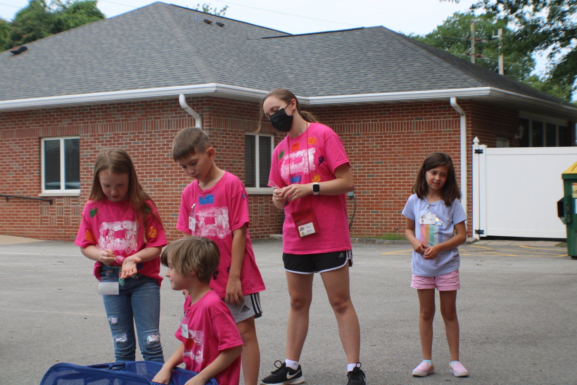 A group of children and a woman wearing pink shirts stand outside a brick building.